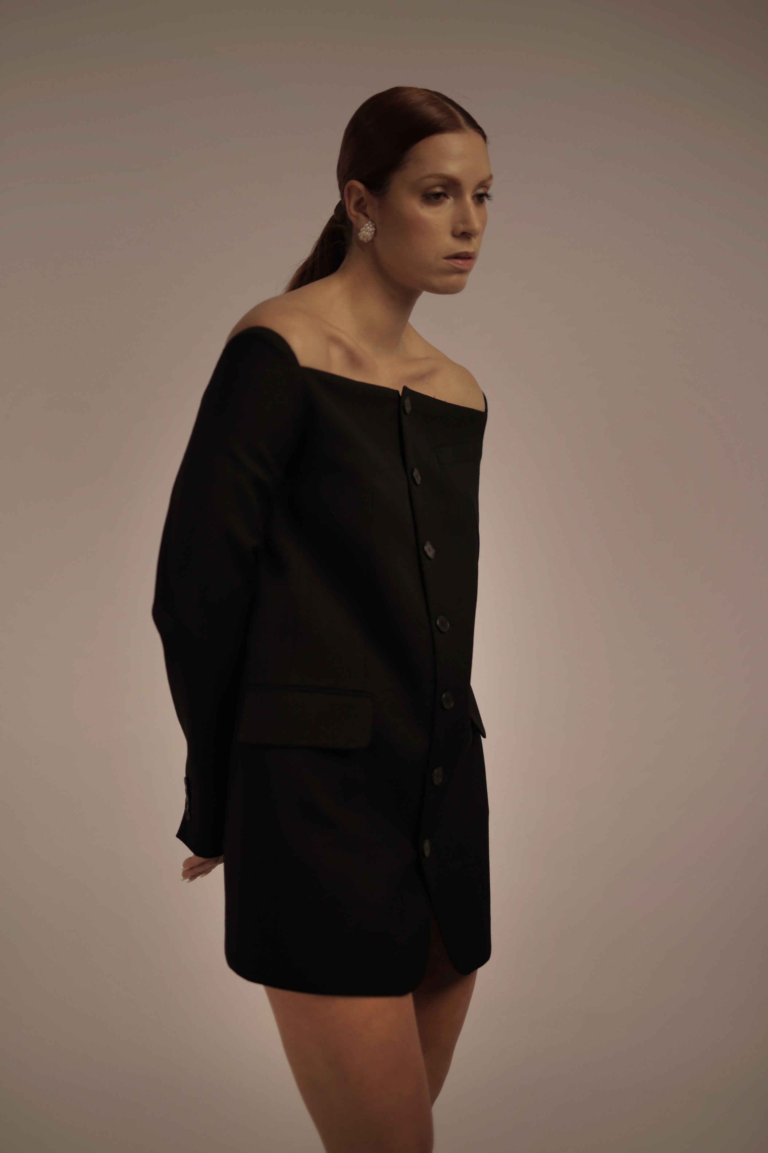 A woman with red hair in a ponytail wearing a black off-the-shoulder blazer dress and pearl earrings stands against a neutral background.