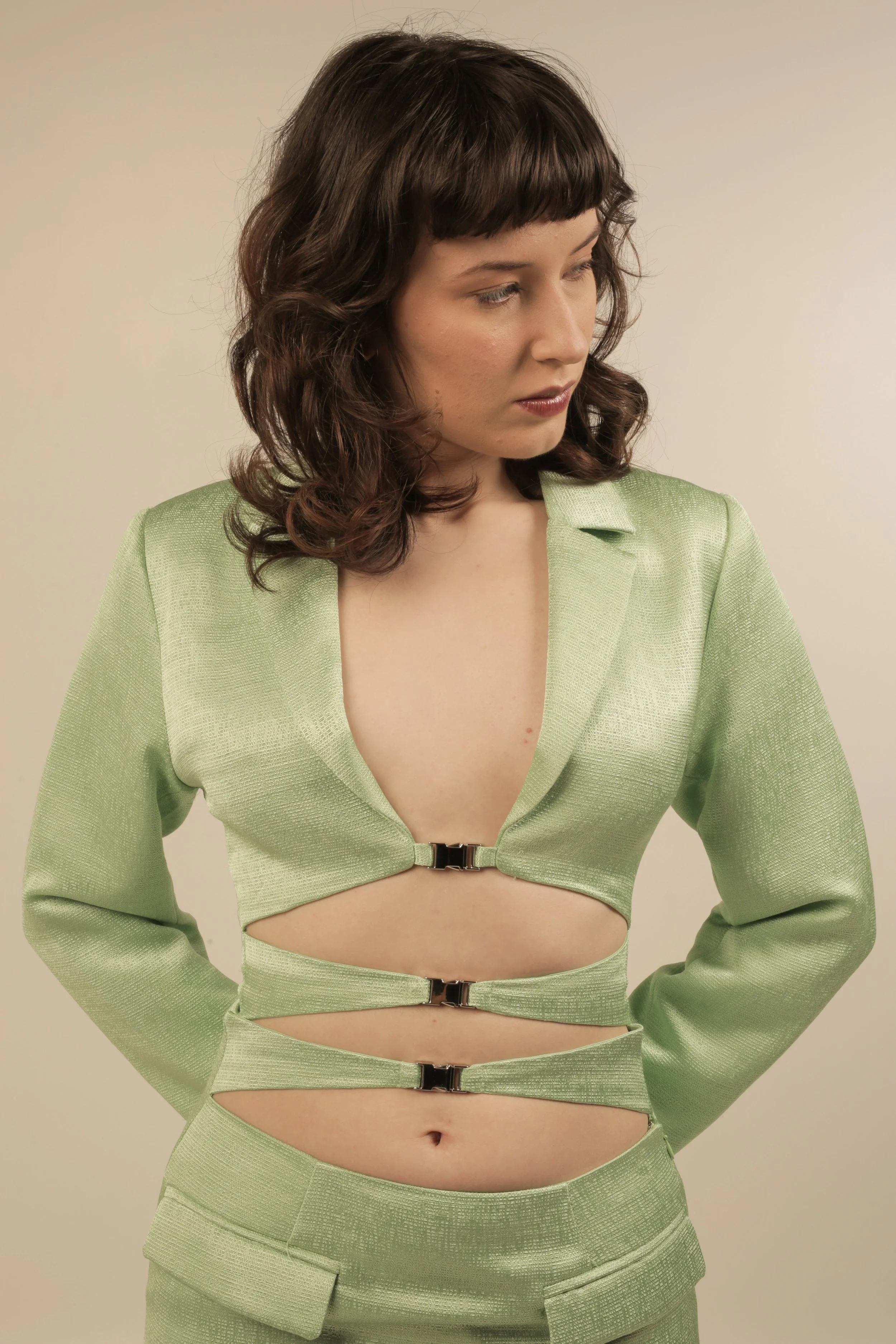 Woman with wavy brown hair wearing a light green, cut-out blazer and matching pants, looking down.