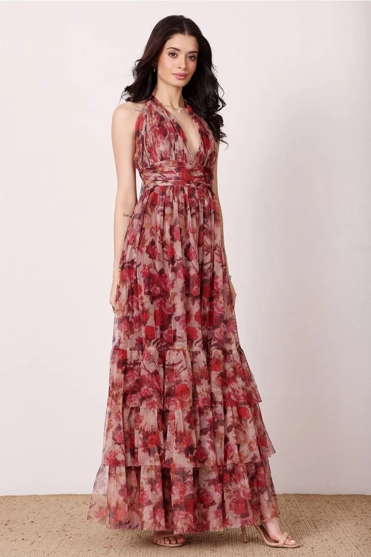 A woman with long dark hair is wearing a sleeveless, floral maxi dress in shades of red and pink, standing on a beige carpet against a plain white wall.