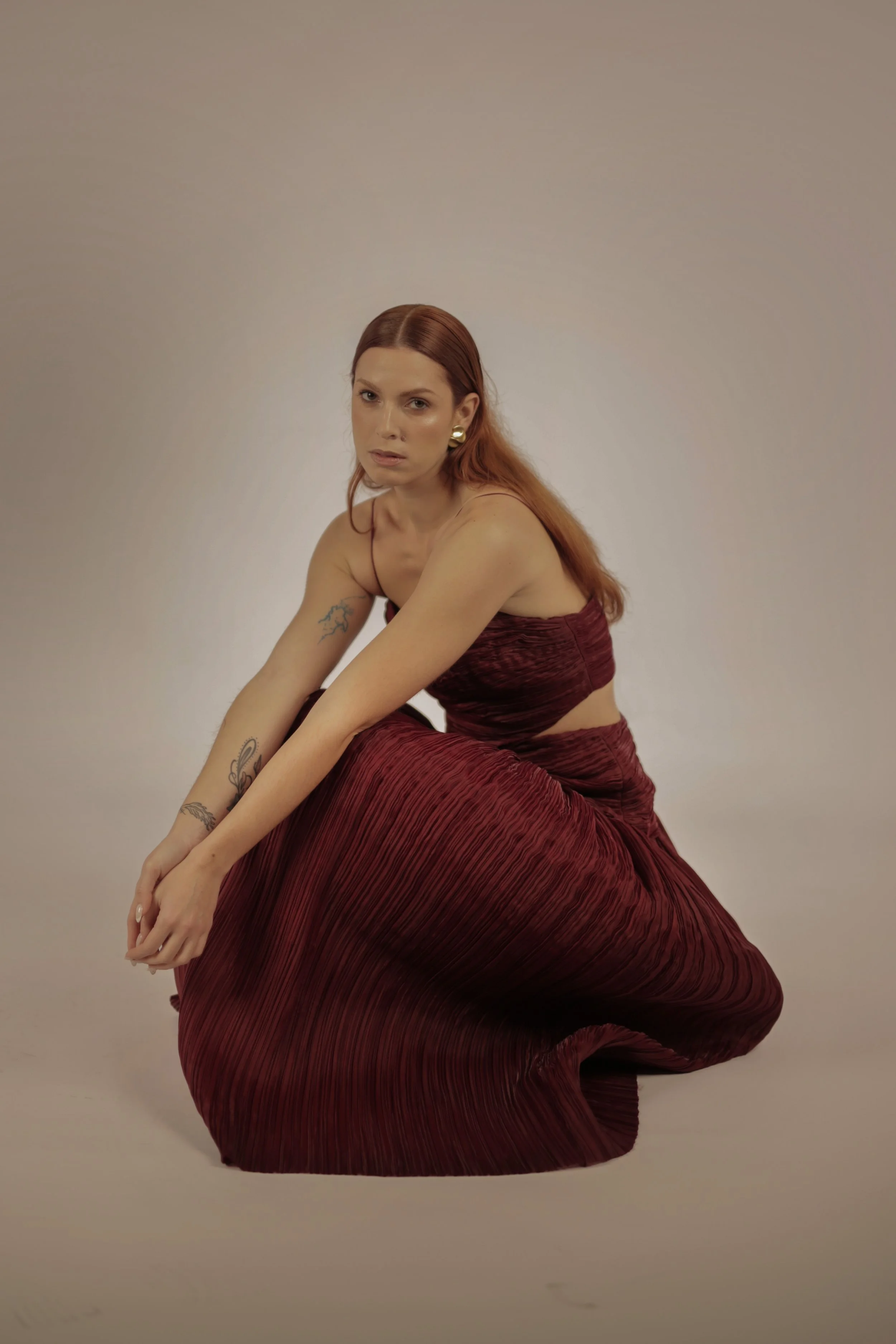 A woman with red hair, gold earrings, and tattoos on her arms, wearing a matching burgundy strapless top and long pleated skirt, poses kneeling against a plain, neutral background.