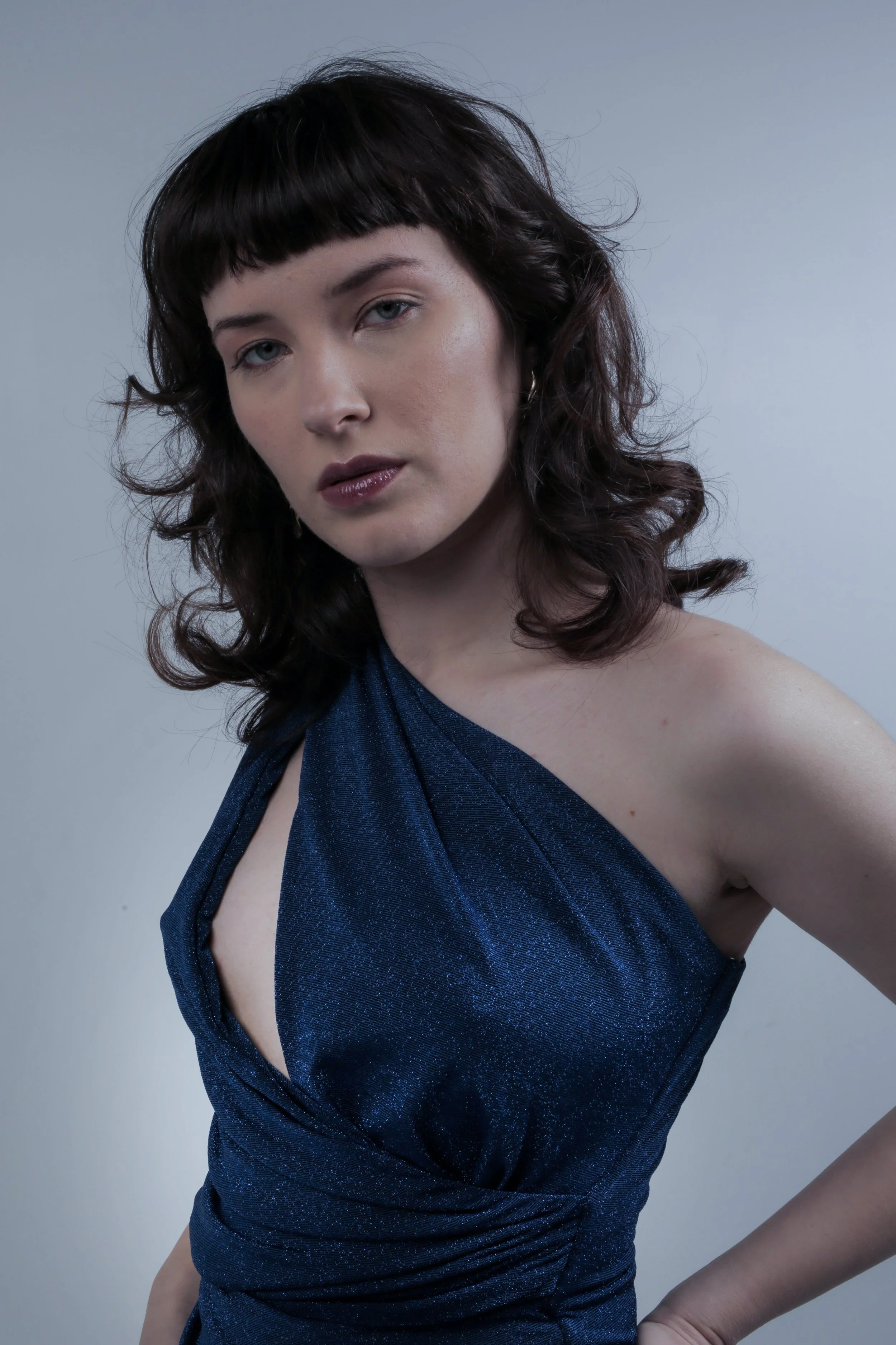 Portrait of a woman with dark curly hair and bangs, wearing a shiny navy blue evening gown with a crowd neckline, against a plain gray background.