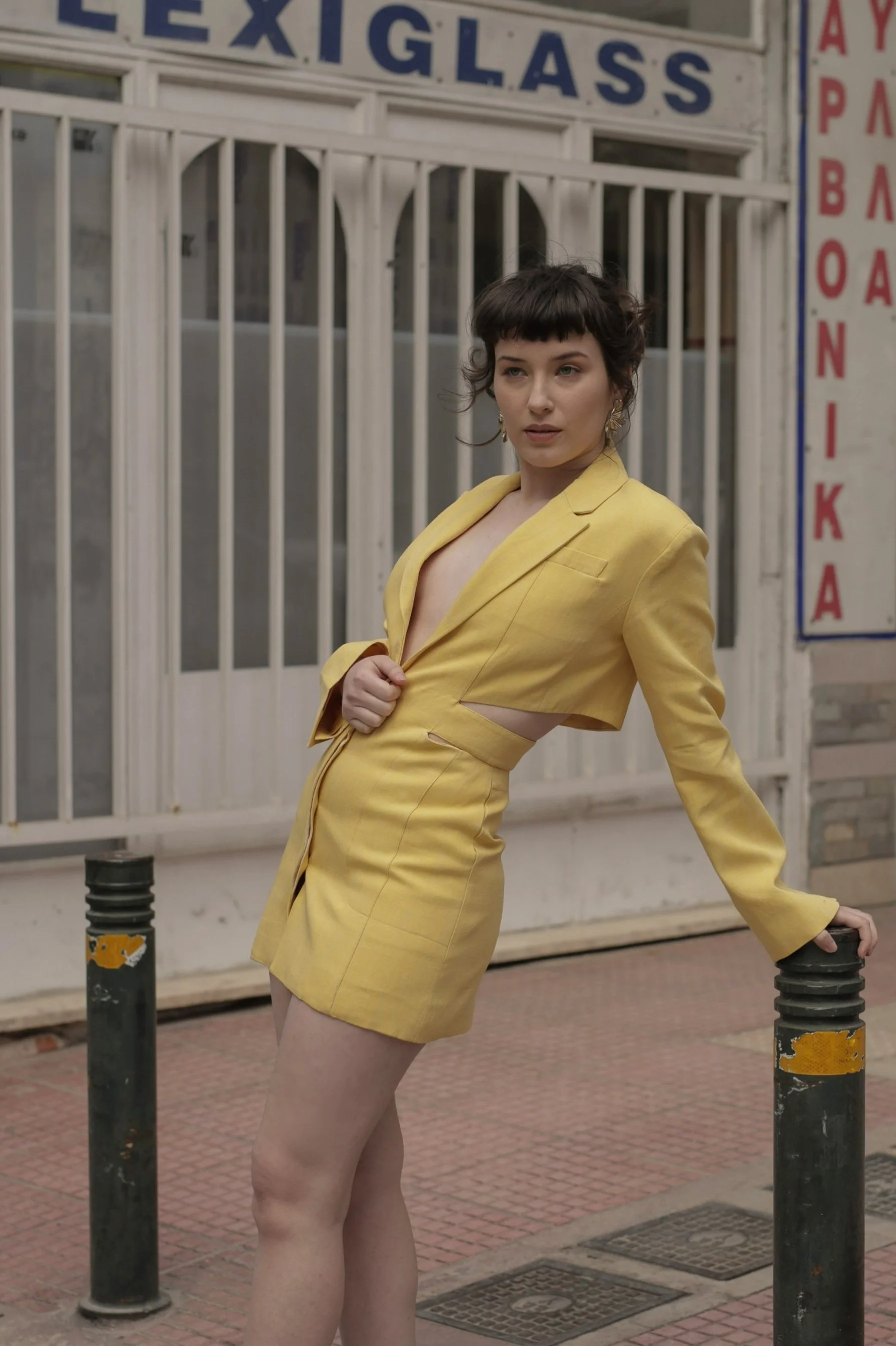 A woman in a yellow blazer and skirt stands outdoors, leaning against black posts, with a building and a sign that says 'LE X GLASS' and other signage in the background.
