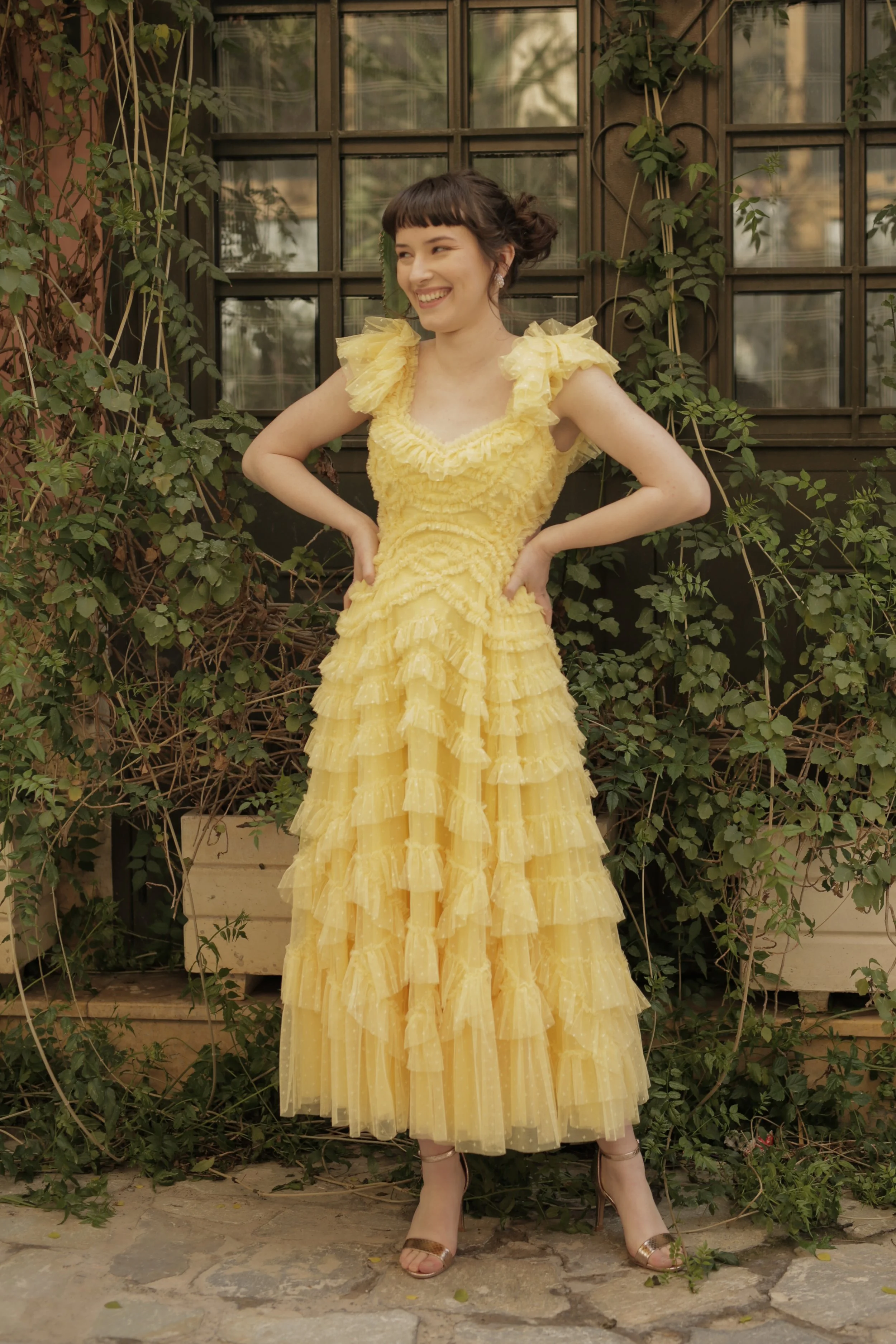 Woman in a yellow ruffled dress smiling with hands on hips standing outdoors in front of a window with greenery.