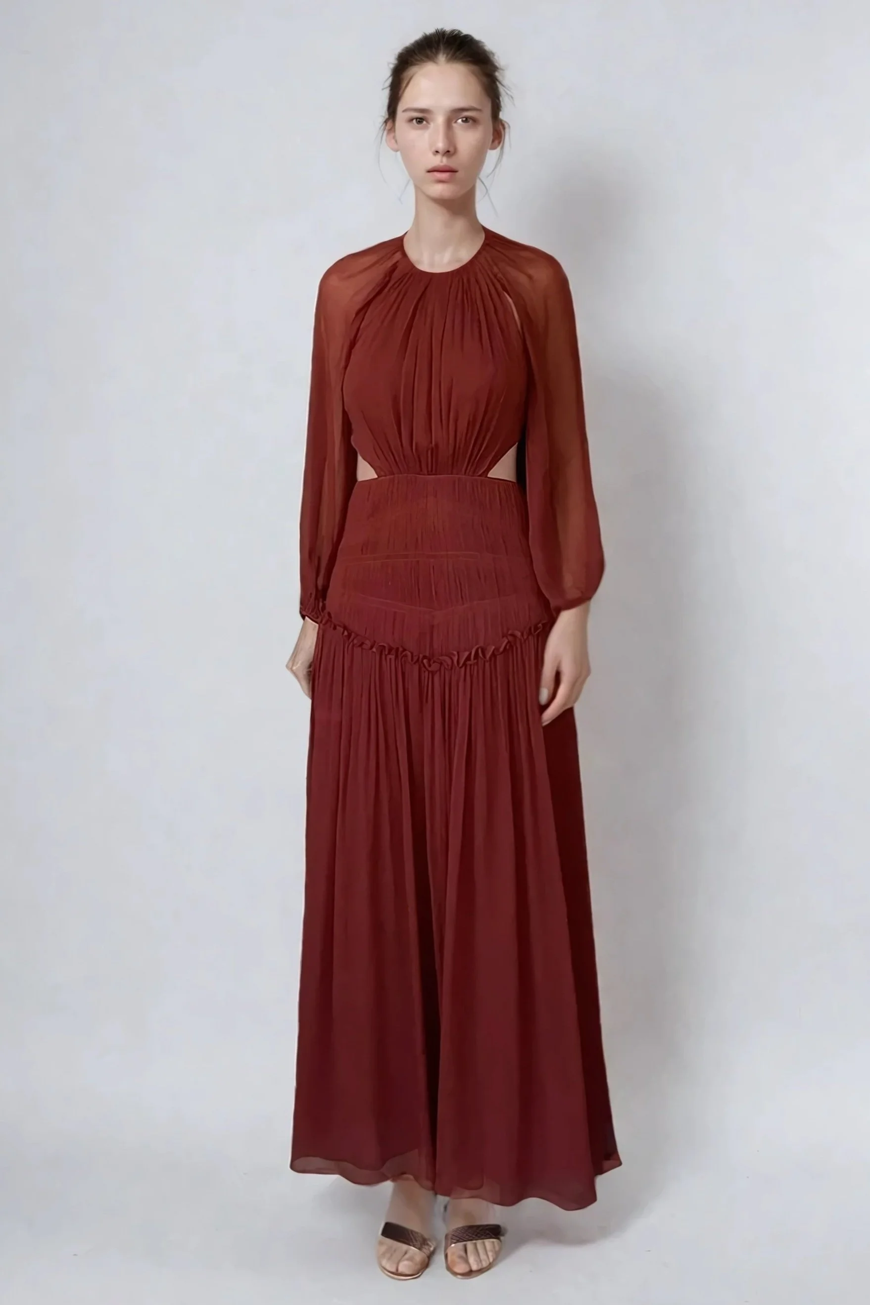 A woman in a long red dress with sheer long sleeves standing against a plain white background.