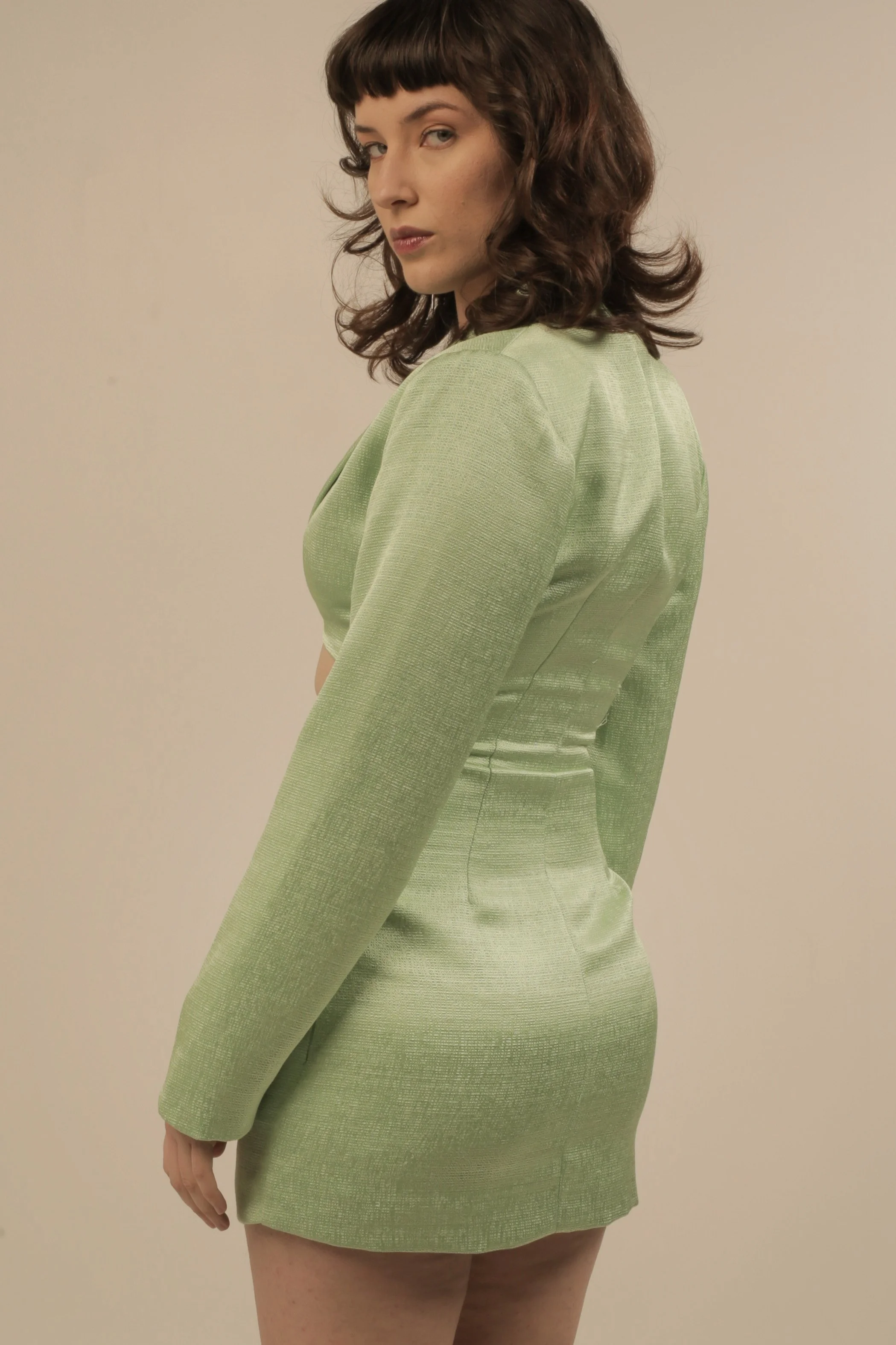 A woman with brown, curly hair and bangs wearing a fitted, green textured dress, standing against a plain beige background, looking over her shoulder.