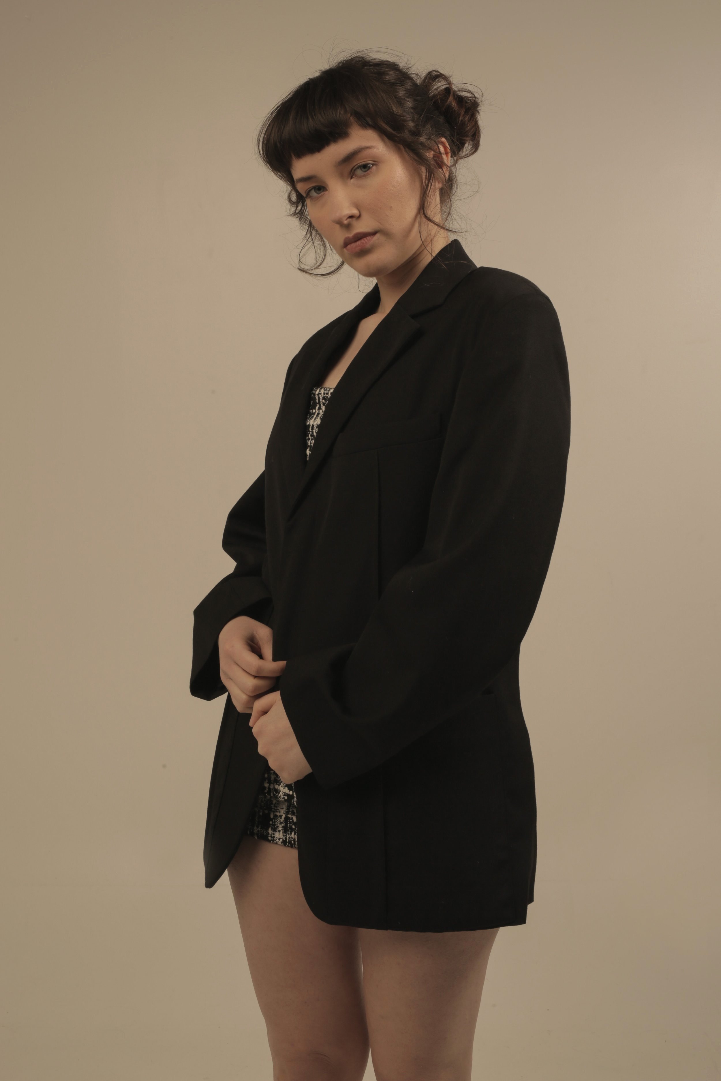 A young woman with dark curly hair and bangs, wearing a black blazer over a patterned dress, standing against a plain beige background.