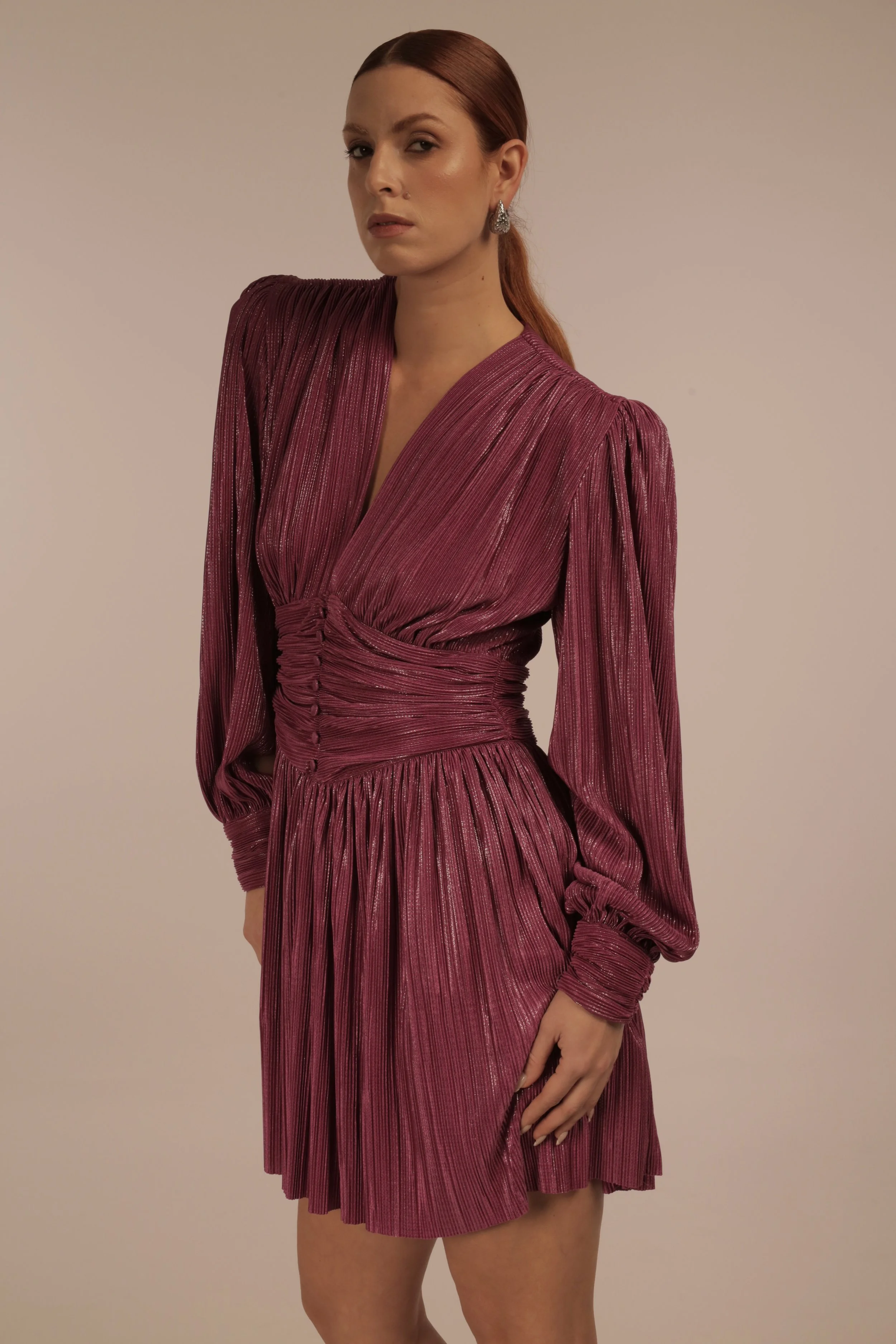 A woman with red hair and earrings wearing a shiny, pleated, deep pink dress with long sleeves and a gathered waist.