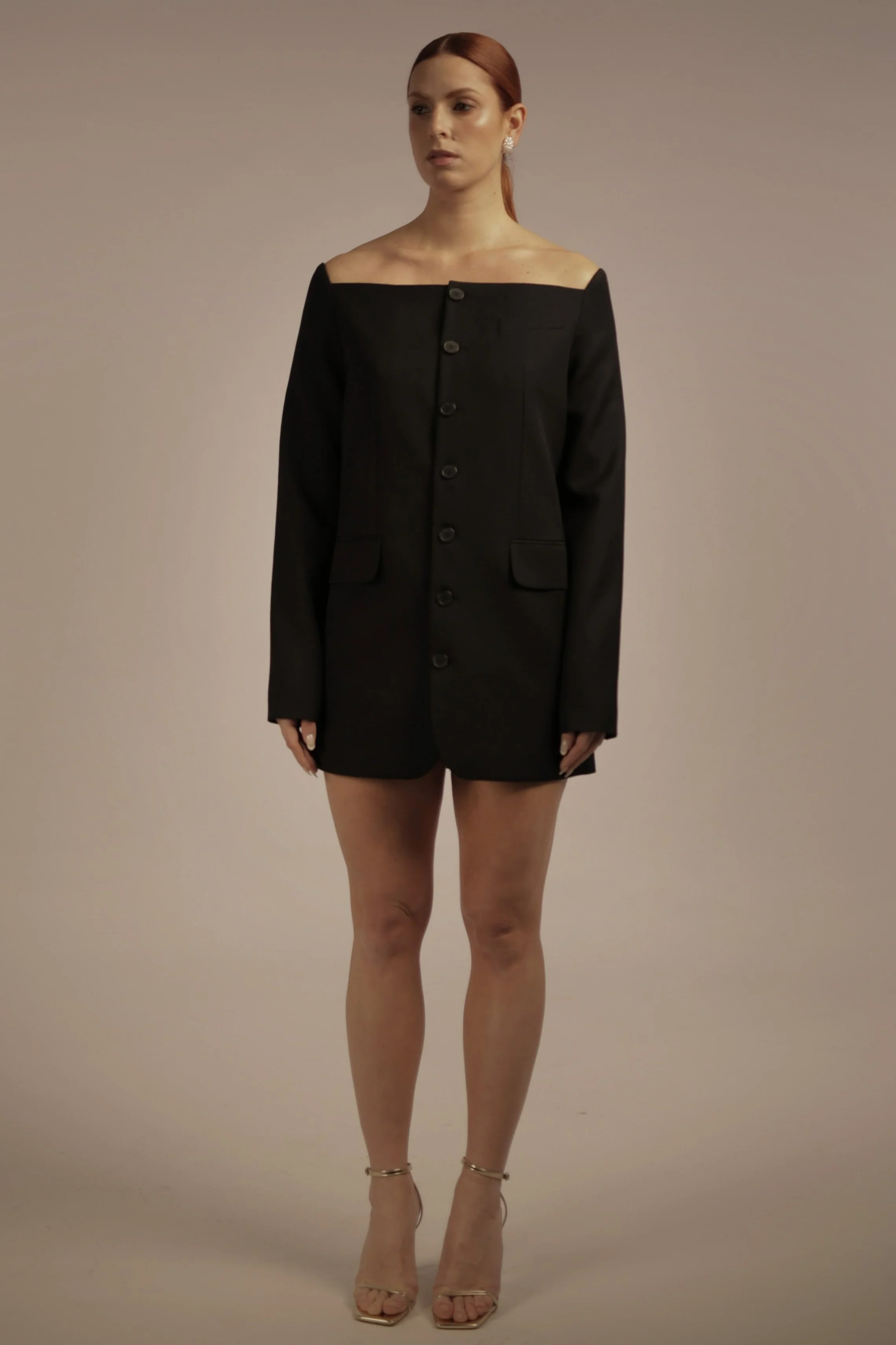 A woman with red hair wearing a black blazer-style dress with long sleeves, a row of buttons down the front, and two front pockets, paired with high-heeled sandals.