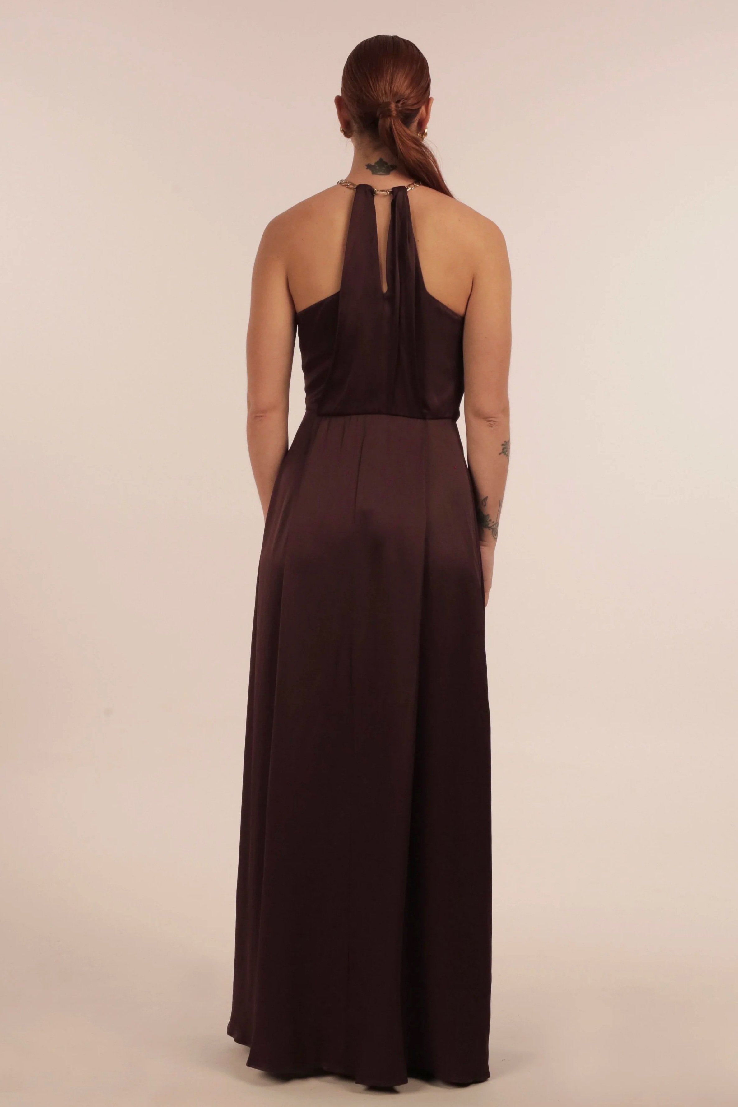 Back view of a woman with red hair in a low ponytail, wearing a dark sleeveless jumpsuit with wide-leg pants. She has tattoos on her right arm and back and is standing against a plain light-colored background.