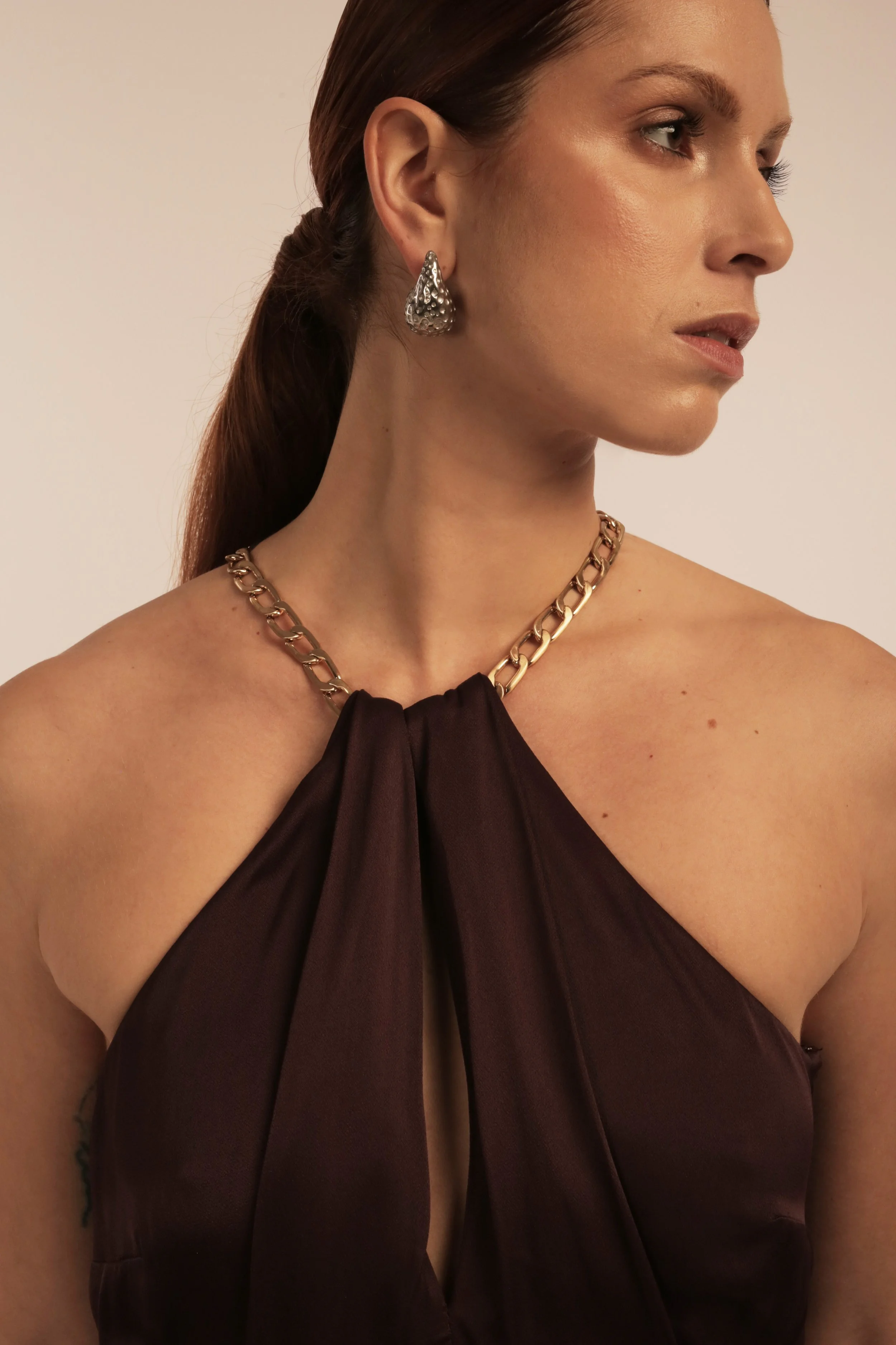 Close-up of a woman with long brown hair, wearing silver earrings and a dark satin halter dress with a gold chain necklace.