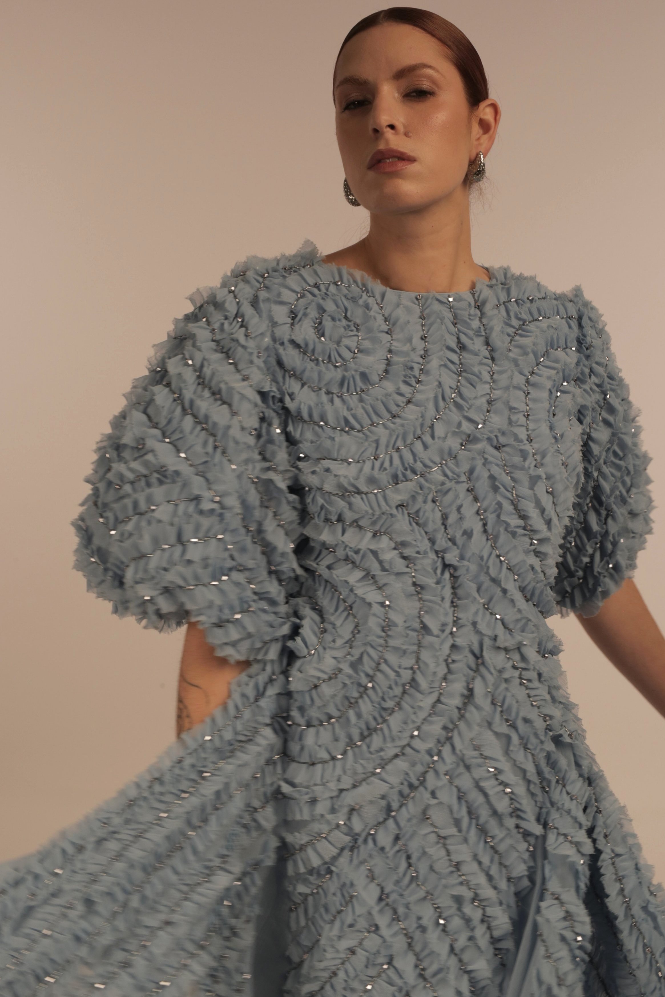 Woman with red hair wearing a textured light blue dress with swirled pattern and metallic accents, standing against a plain background.