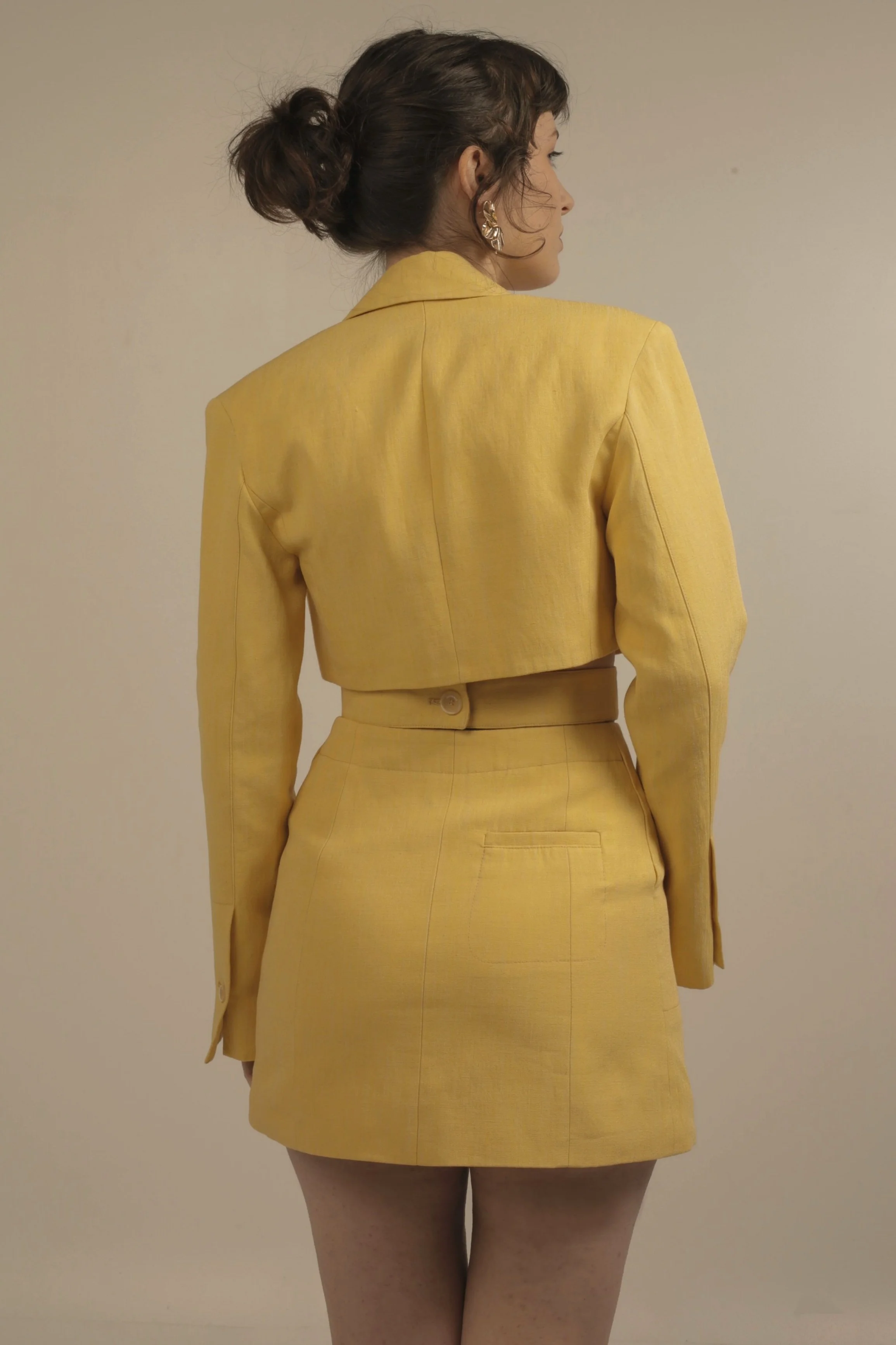 A woman with dark hair tied in a messy bun, wearing gold earrings and a yellow two-piece suit with a cropped blazer and matching skirt, standing against a plain beige background.