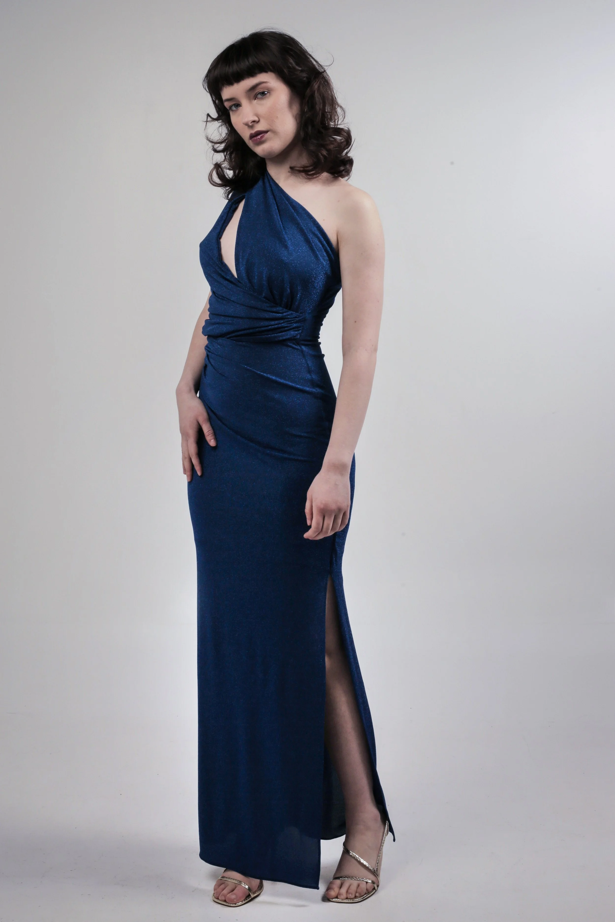 A woman with dark hair and bangs wearing a blue, one-shoulder evening gown with a slit, standing against a plain background.
