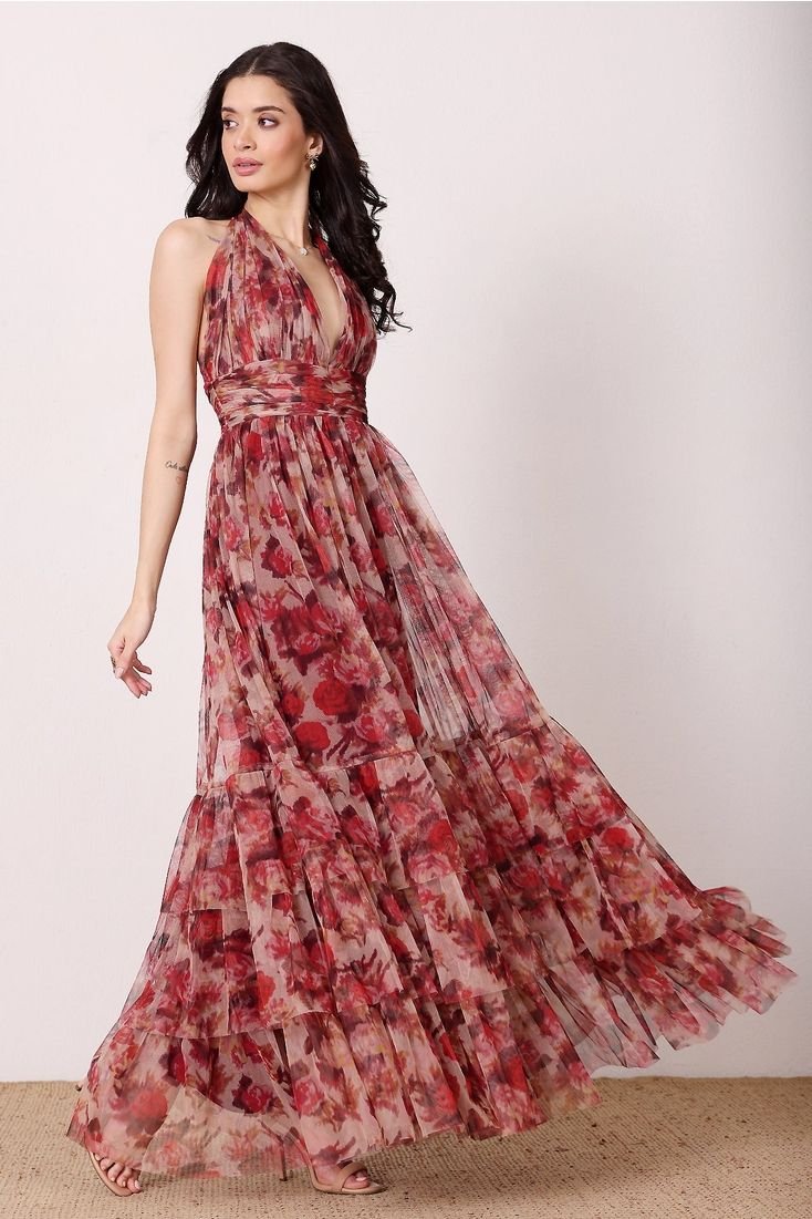 A woman in a long, flowing floral dress with shades of red, pink, and brown standing against a plain white background.