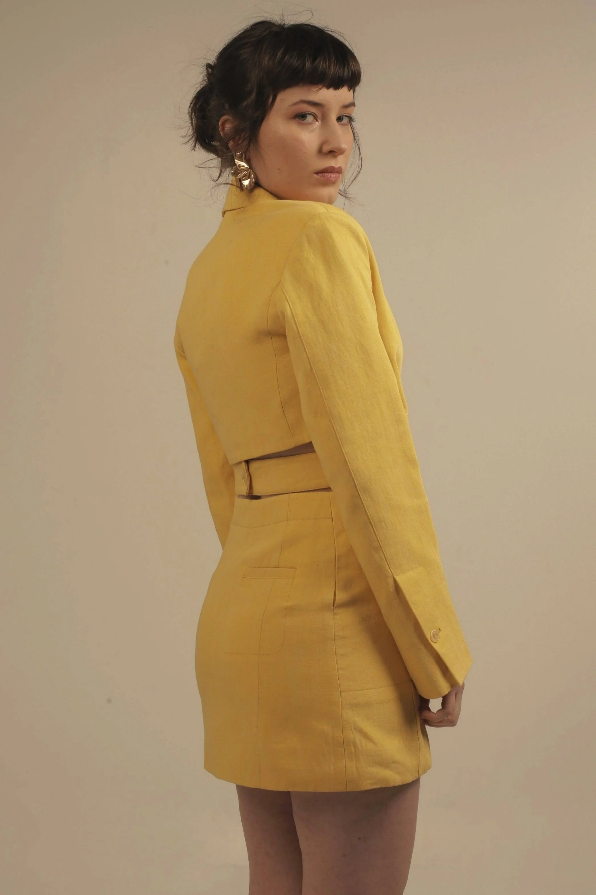 A woman with short dark hair in a yellow suit, standing against a beige wall, looking over her shoulder.