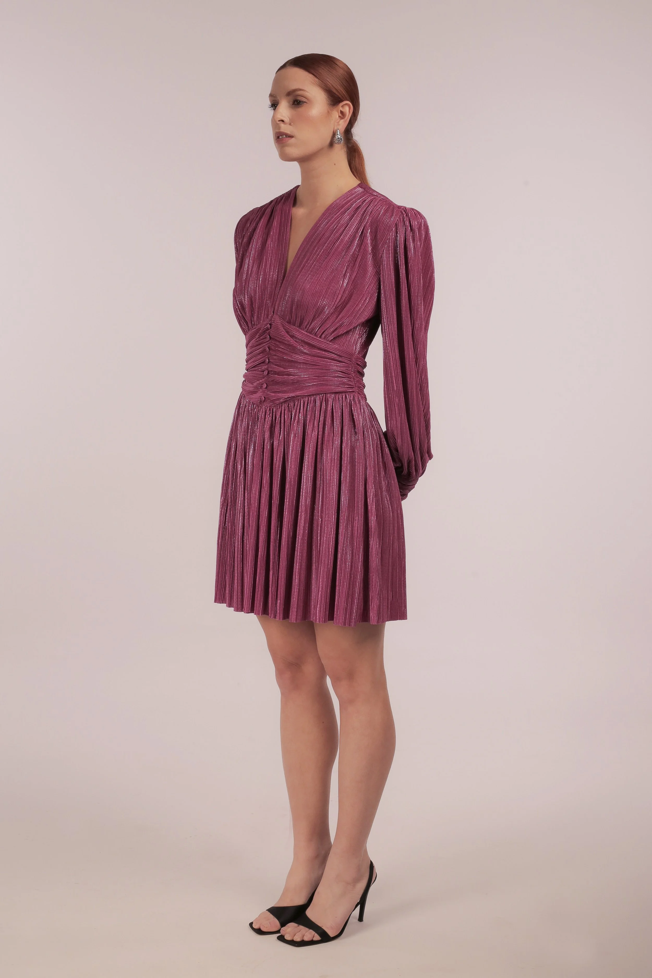 Woman with red hair wearing a shiny, pleated pink dress with long sleeves and black high heels.