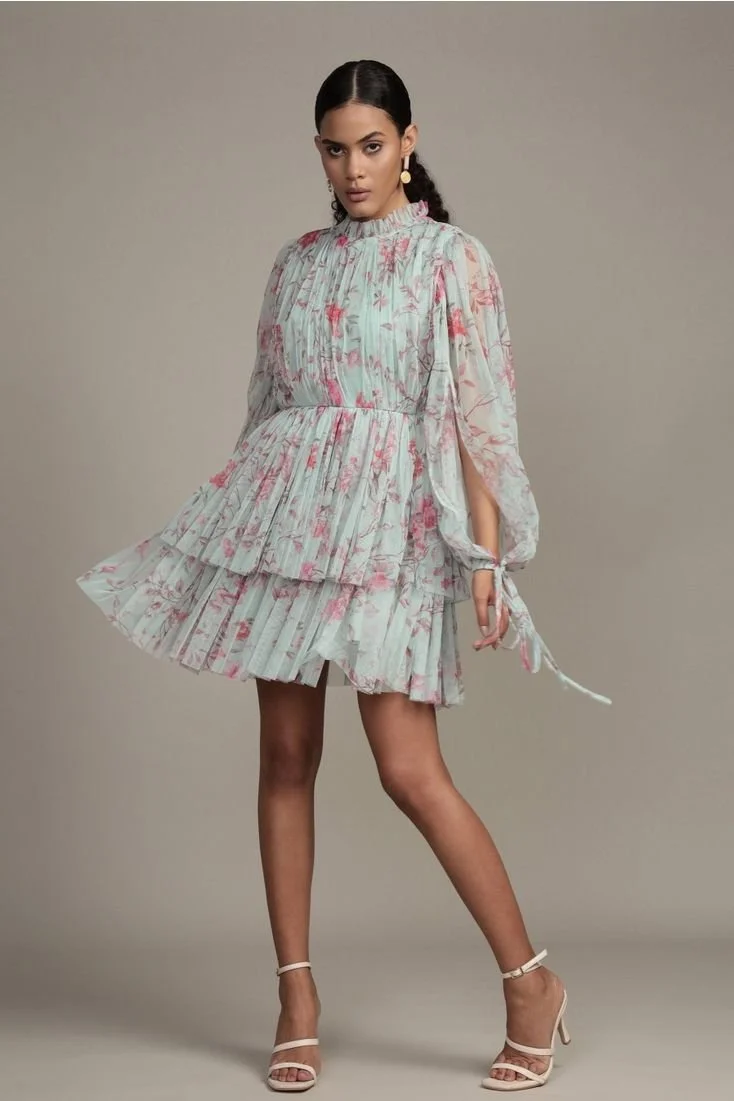 Woman wearing a floral printed, pleated dress with layered skirt, accessorized with dangling earrings, standing with one leg slightly forward against a plain background.