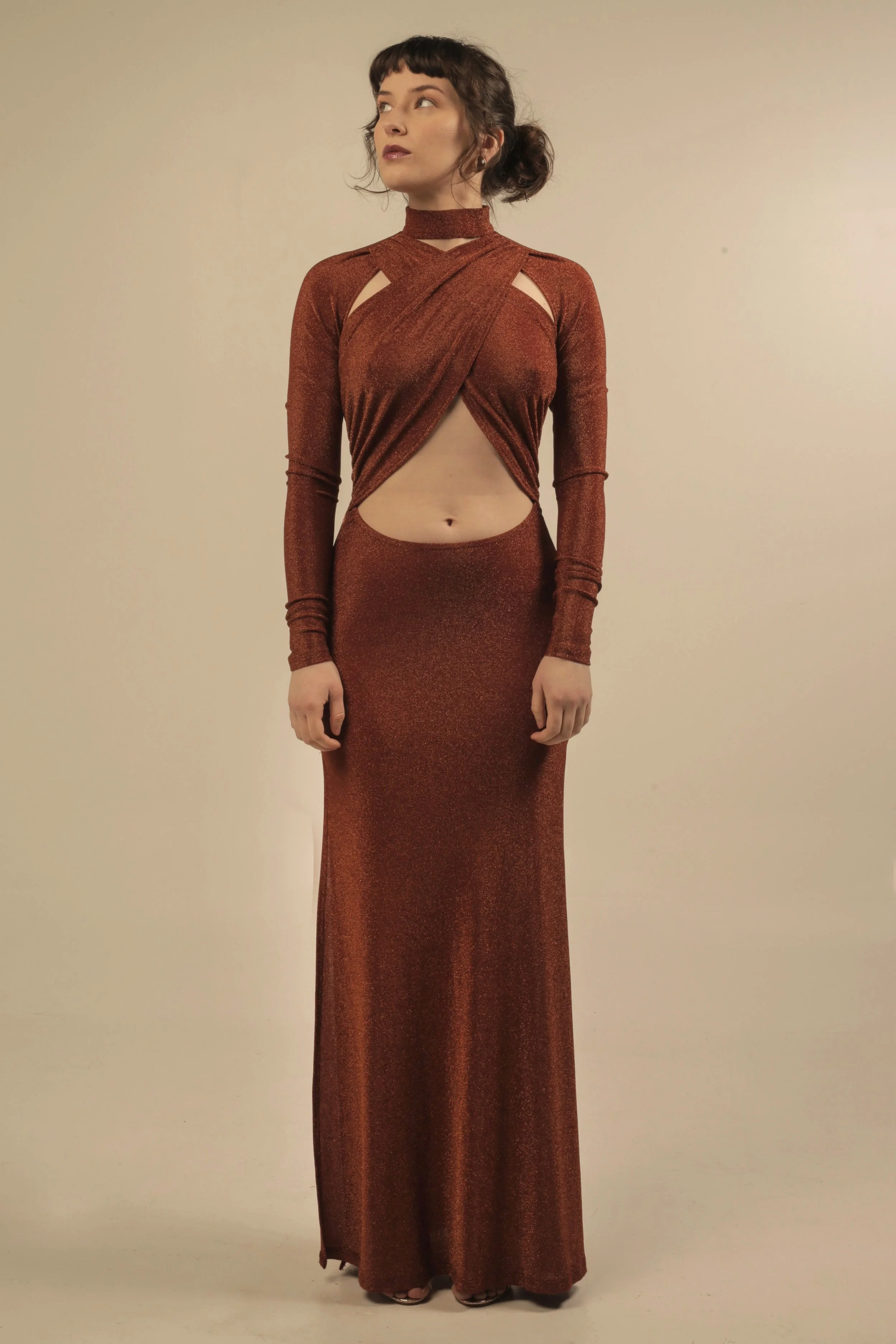 Young woman in a shimmering brown, cut-out, floor-length gown standing against a plain background.