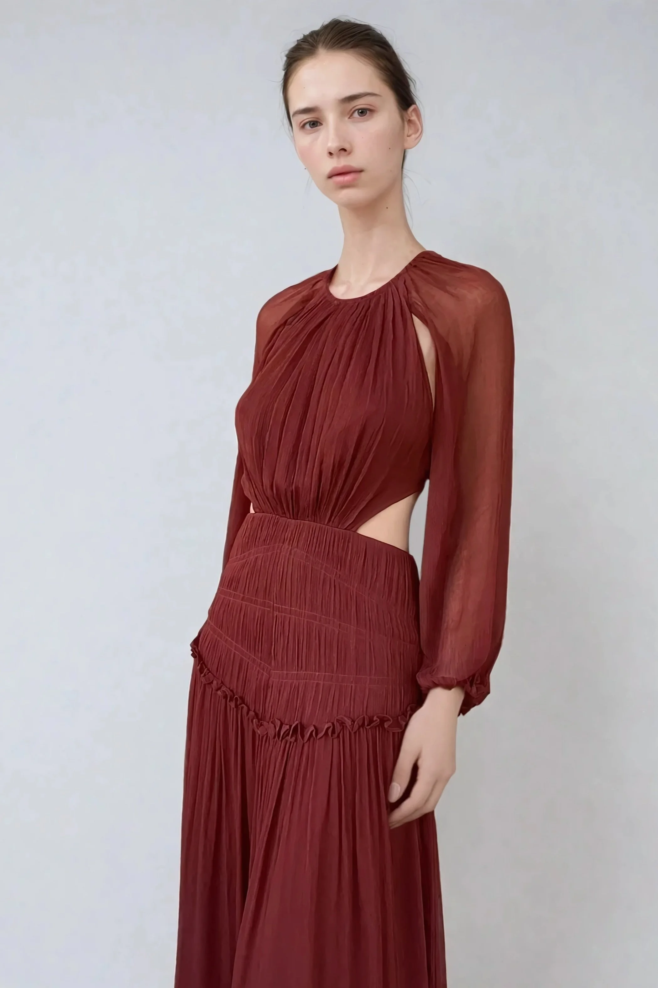 A young woman with brown hair pulled back wearing a long reddish-brown pleated dress with long sheer sleeves and cut-out sides.
