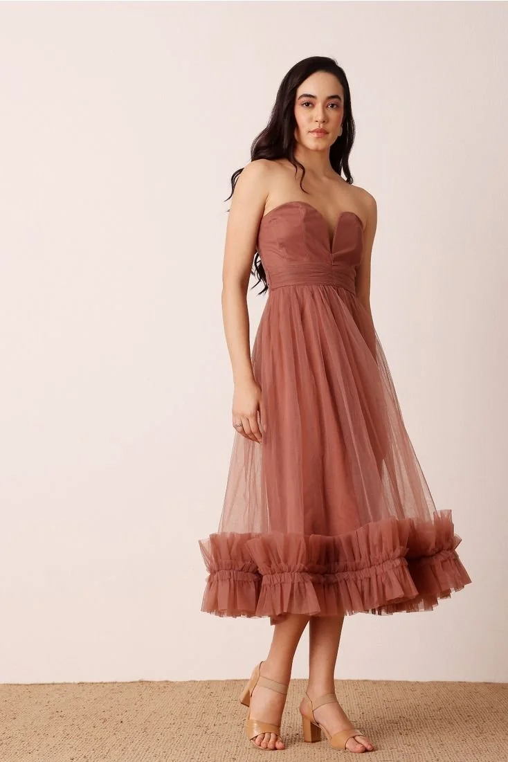 A woman with long dark hair wearing a strapless, dusty rose satin and tulle midi dress with ruffled hem, standing against a plain off-white background.