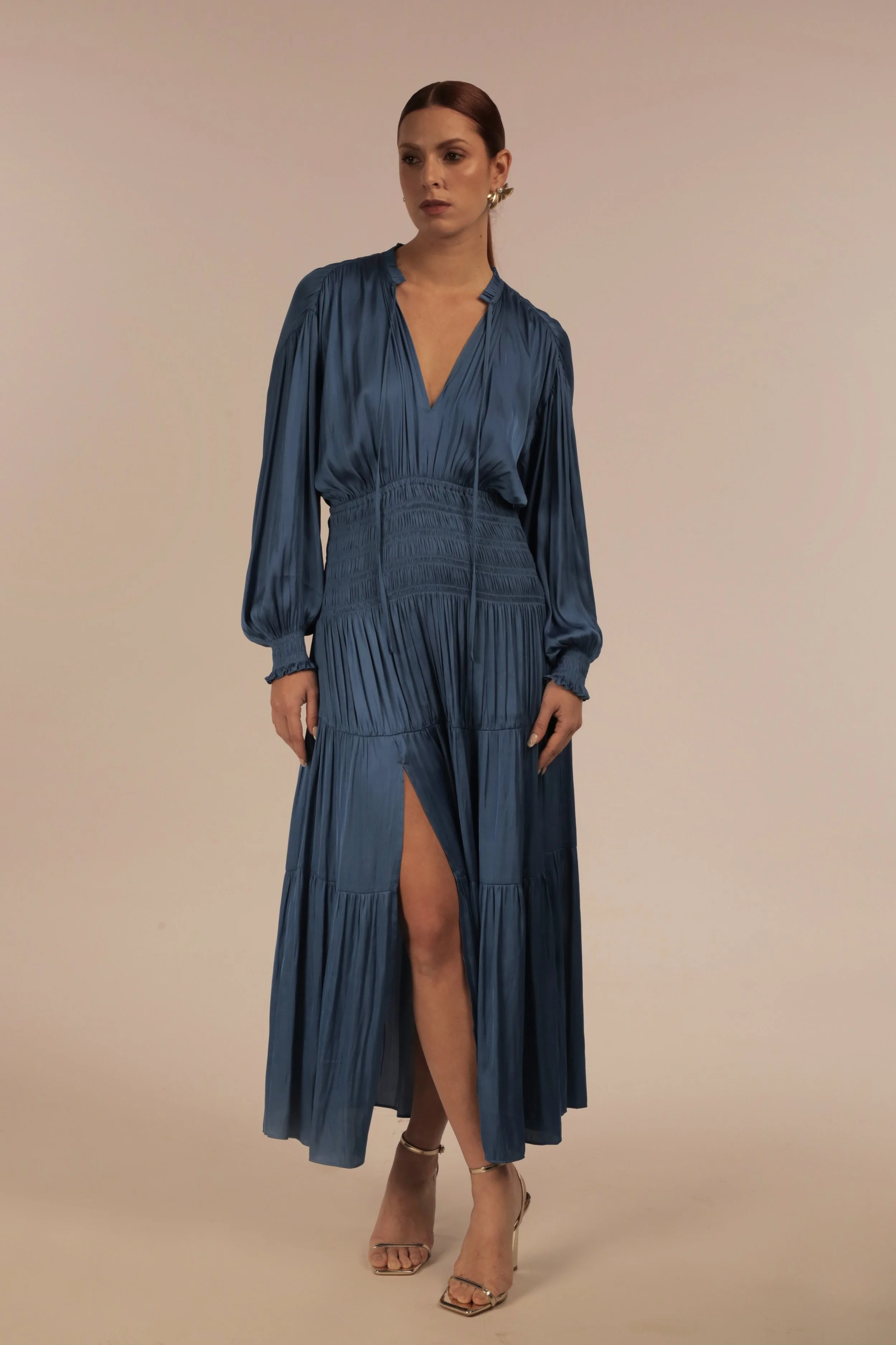 A woman in a long, shiny blue dress with a thigh-high slit, standing against a plain, beige background.