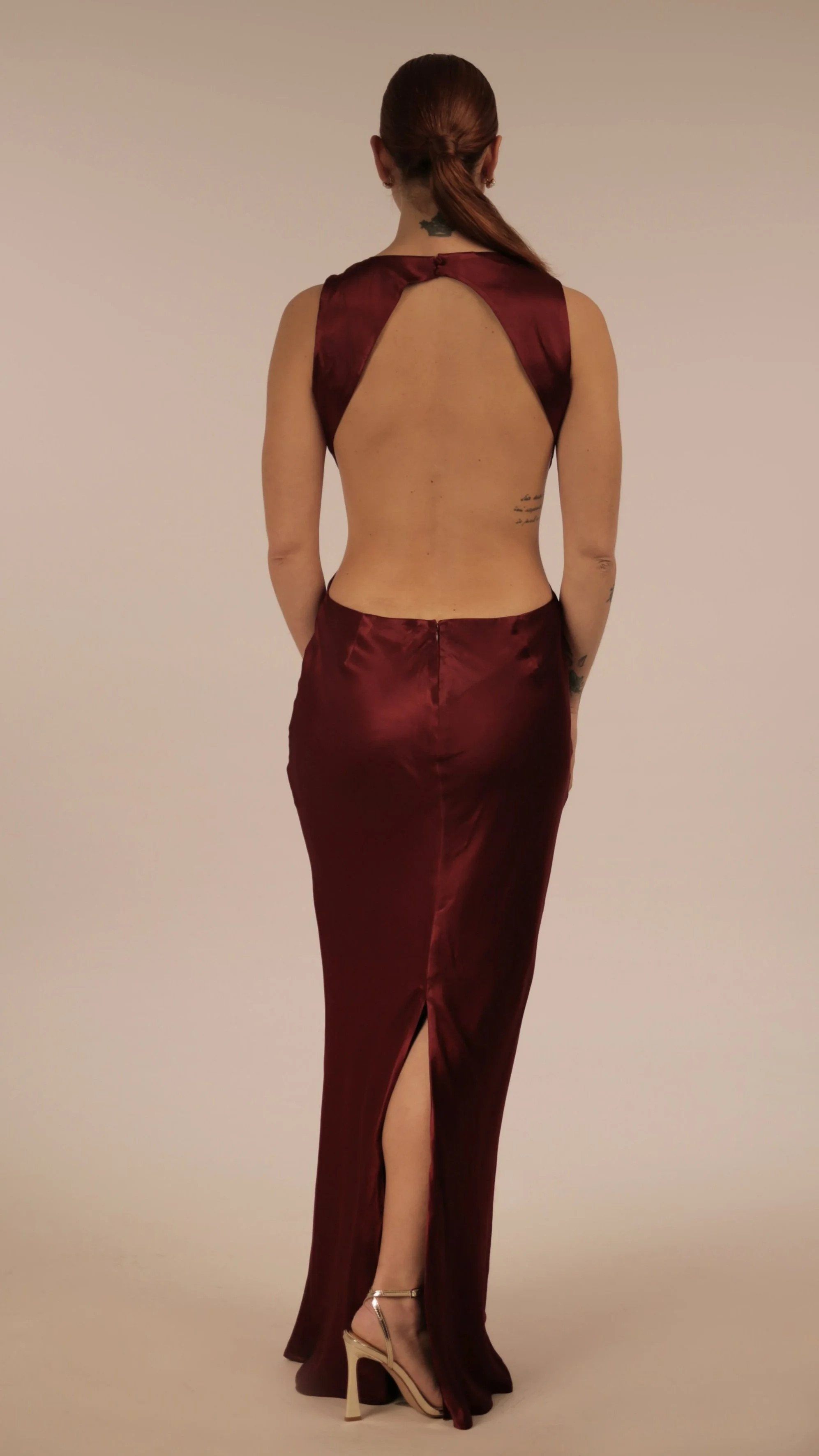 A woman in a burgundy satin dress, viewed from the back, with a high slit and open back design, wearing heels, standing against a neutral background.