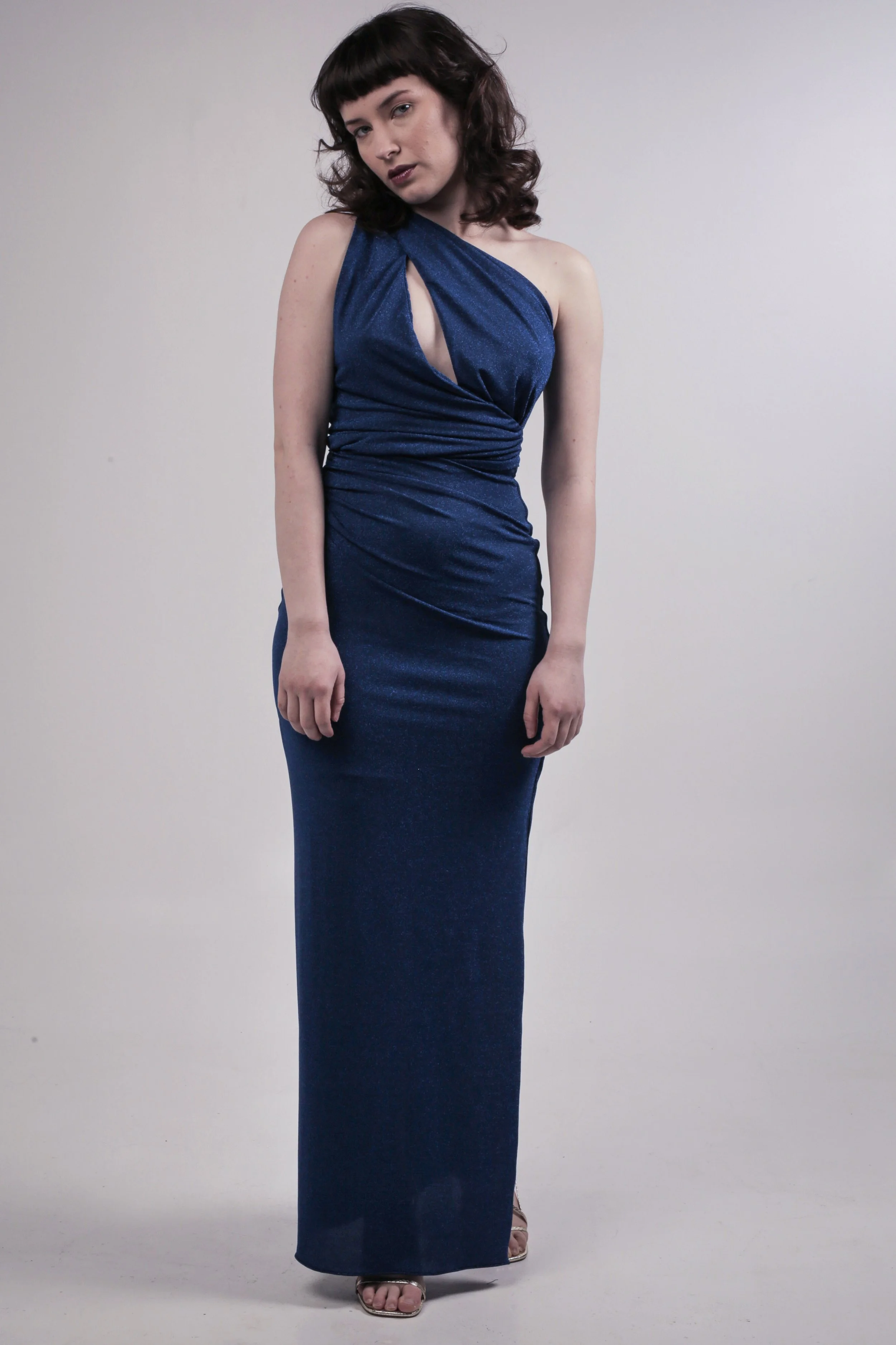 A woman in a dark blue, one-shoulder, floor-length dress with a cutout detail on the chest, standing against a plain light background.