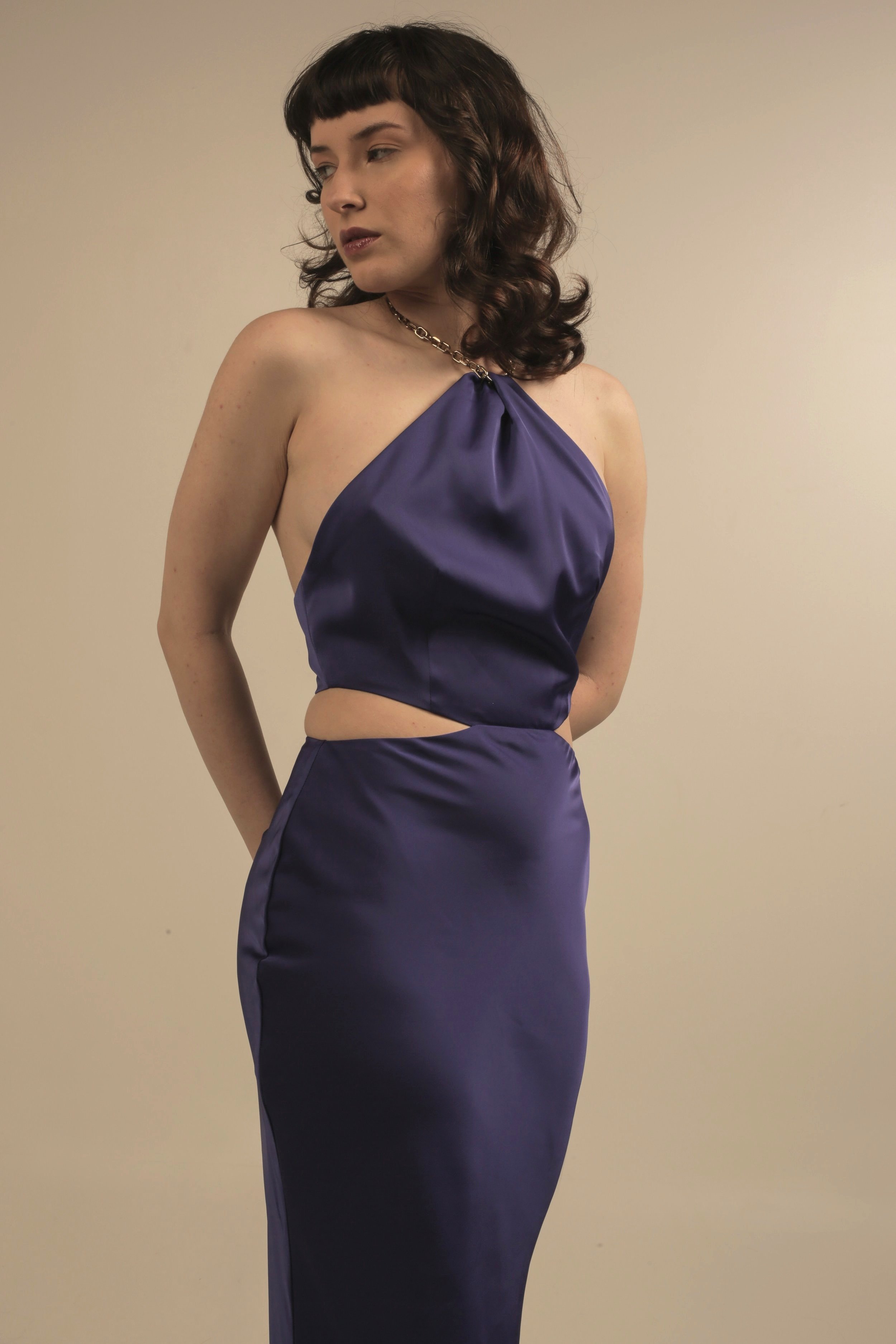 A woman with dark brown, curly hair and bangs, wearing a purple satin dress with a halter top and cutout at the waist, standing against a plain light-colored background.