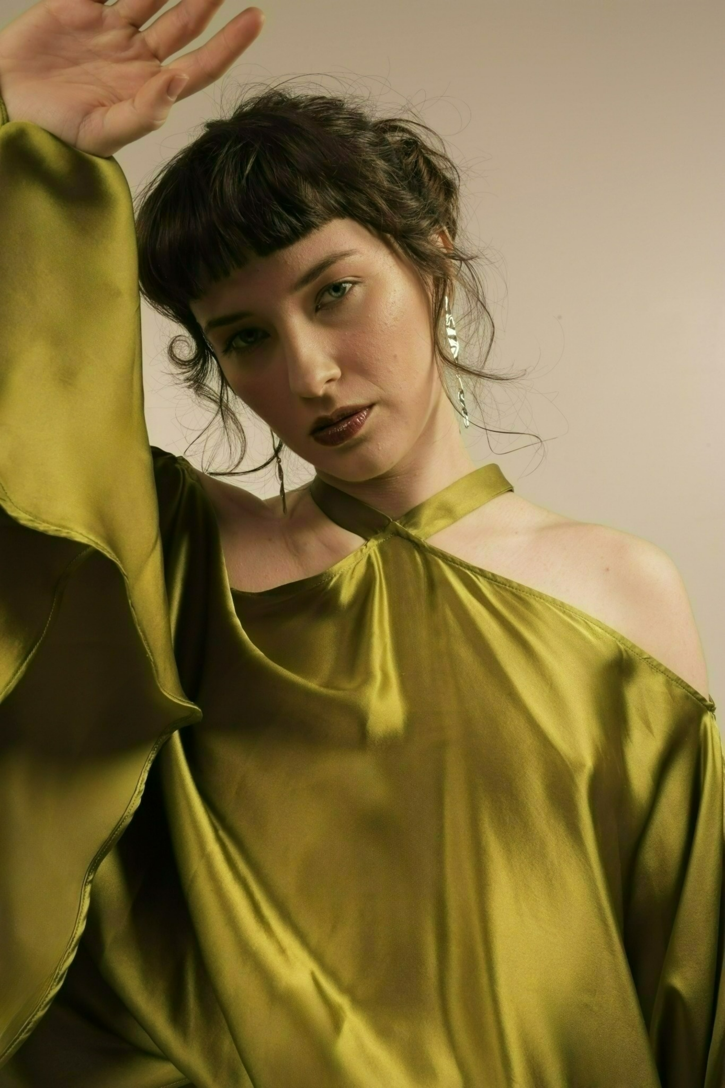 A woman with short dark hair and bangs, wearing dark lipstick, a shiny gold silk off-shoulder dress, and large silver earrings, posing with her hand raised near her head against a plain background.