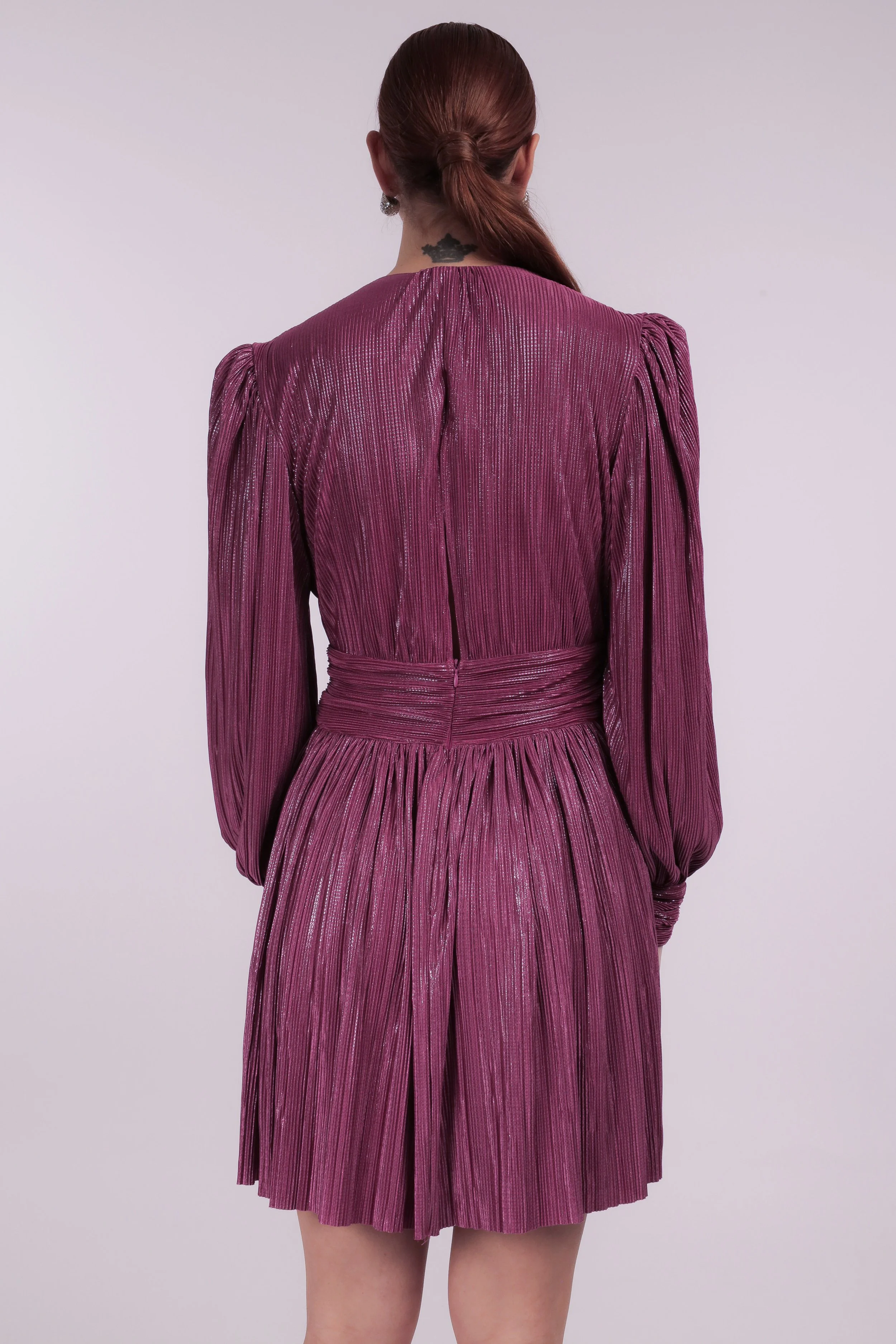 Back view of a woman with reddish-brown hair tied in a low ponytail, wearing a shiny, pleated magenta dress with long sleeves and a high waist, against a plain light purple background.