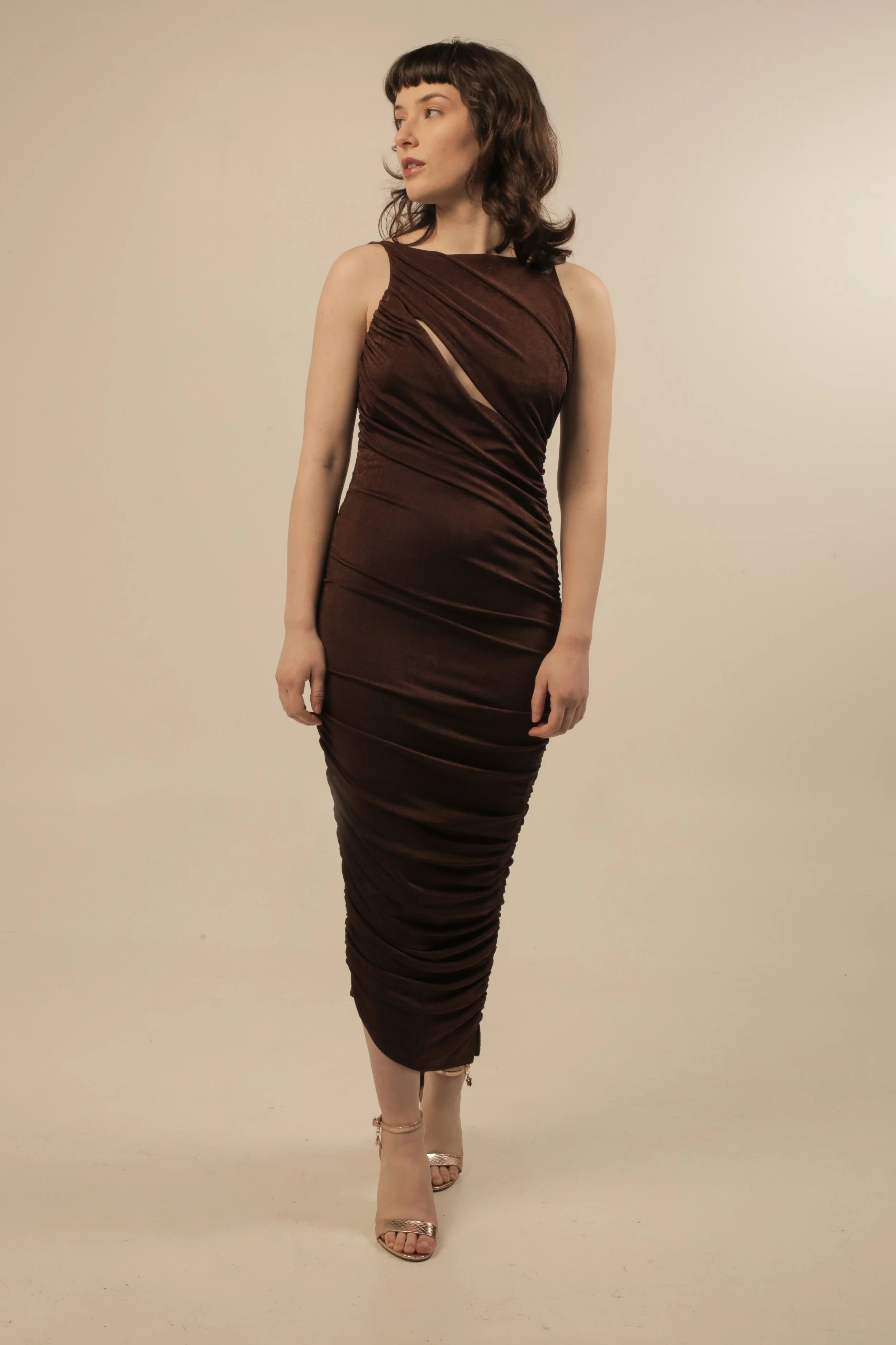 A woman with short dark hair wearing a fitted, sleeveless, dark brown dress with textured detail, standing against a plain light-colored background.