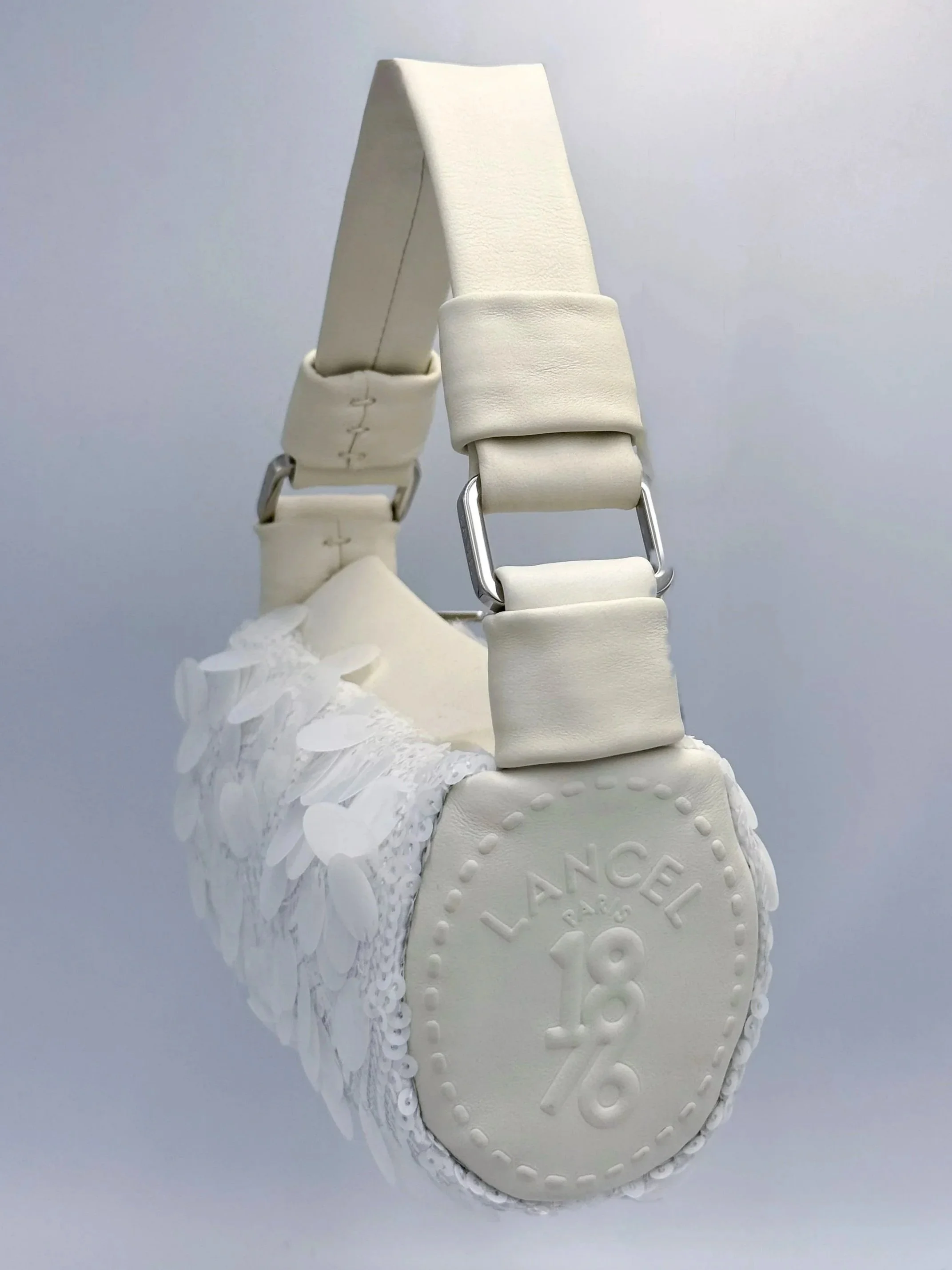 A designer white sneaker with floral embellishments and a strap across the ankle. The sole has a circular detail with the brand name 'Lance Paris' and the number '18/76' embossed on the bottom.