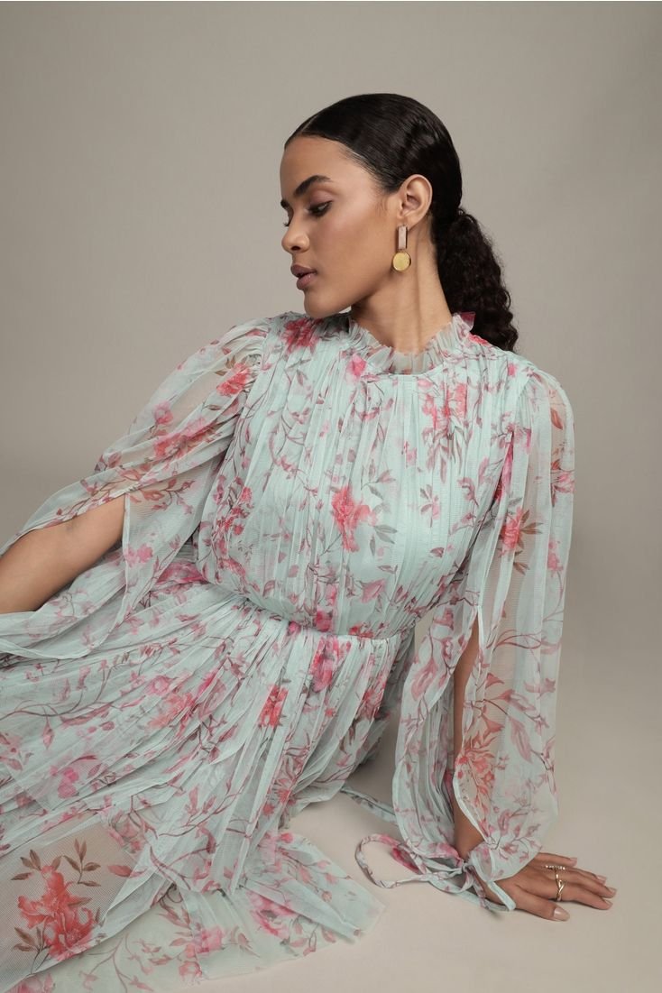 A woman with dark, curly hair and earrings wearing a light, floral, flowing dress with pink and peach patterns, sitting against a plain, neutral background.