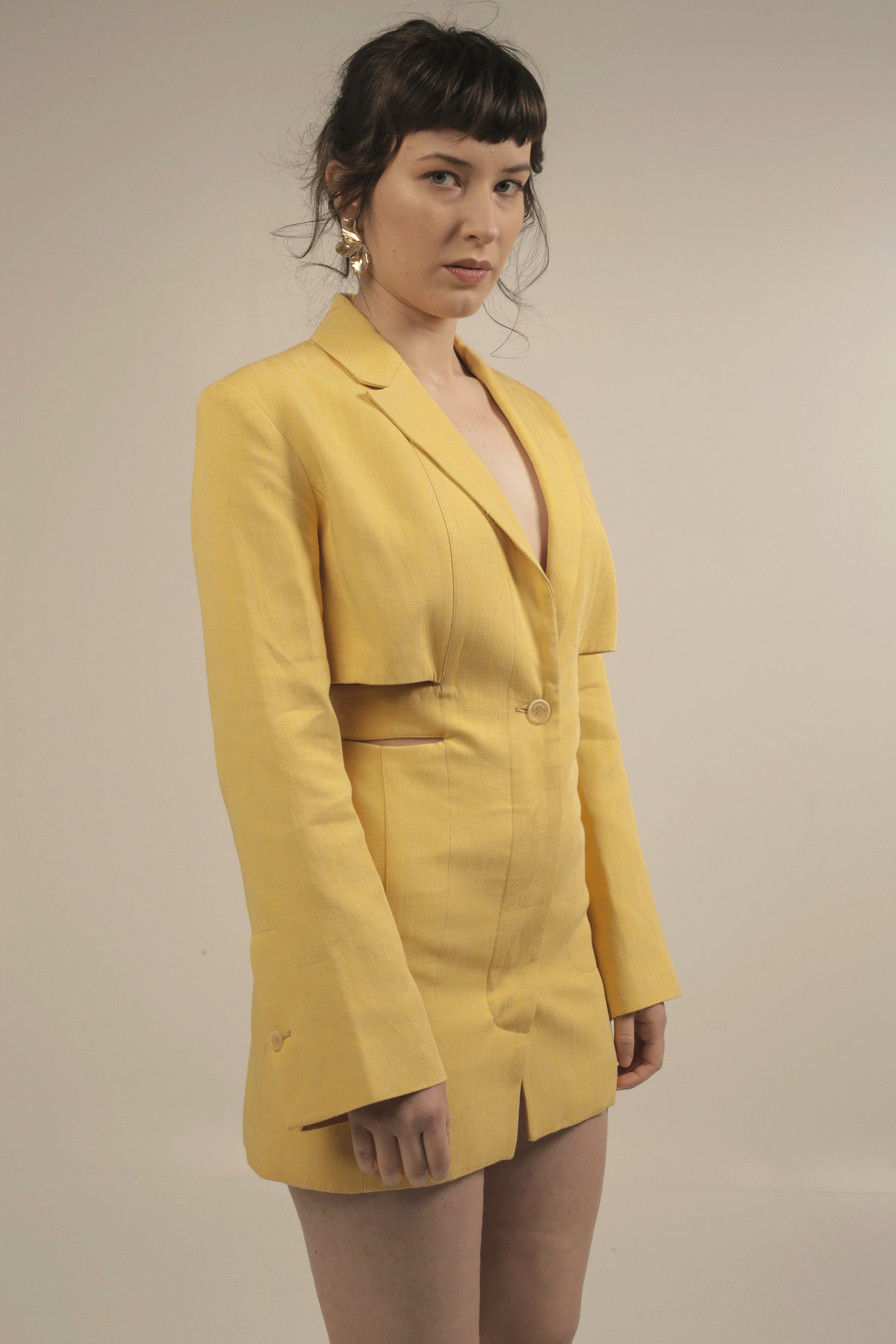 A woman with dark hair in a short haircut, wearing a yellow blazer and earrings, standing against a plain background.