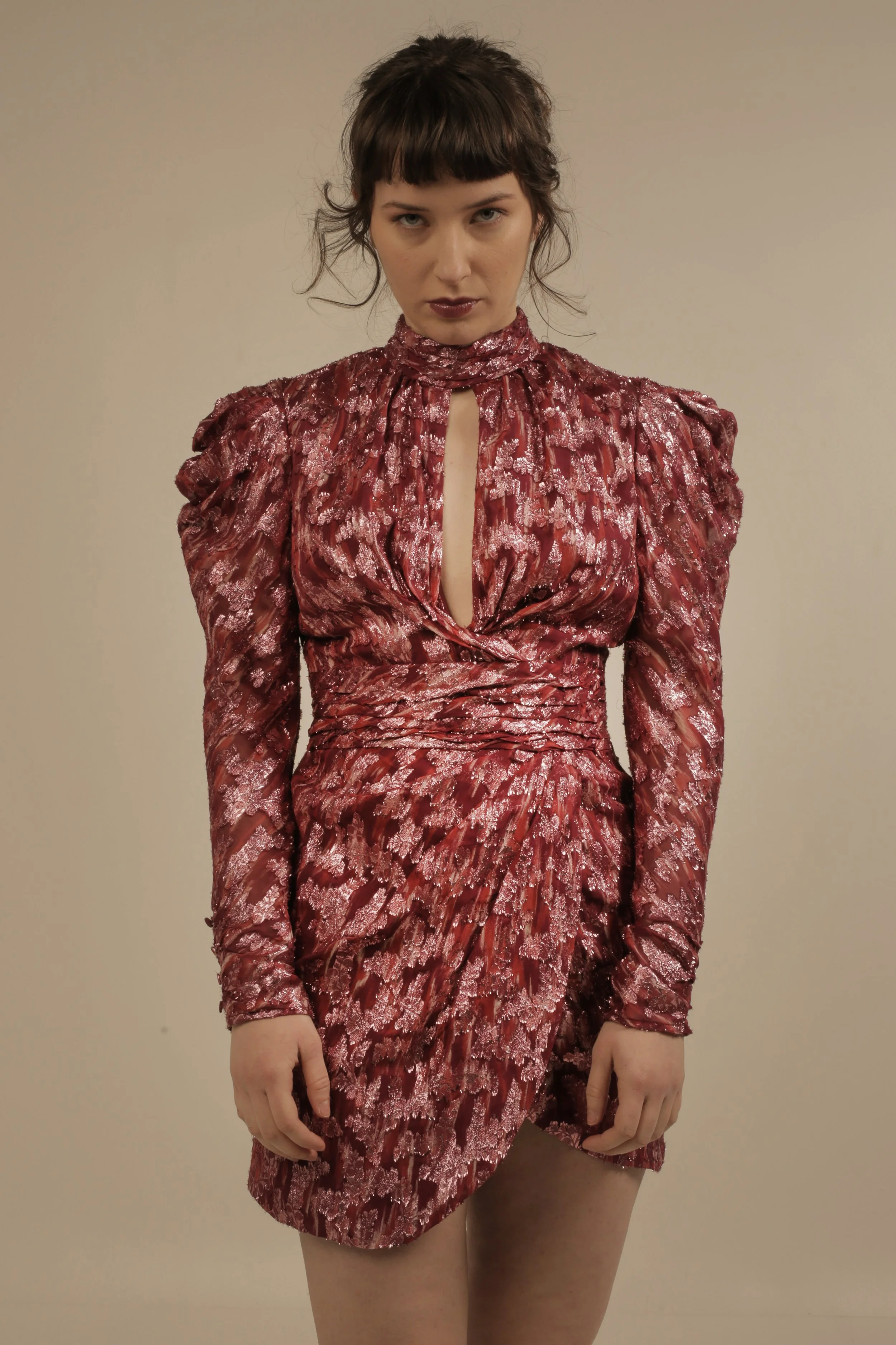 A woman with dark hair, styled with bangs, wearing a shiny, textured red dress with long sleeves, a high collar, and a keyhole detail in the front, standing against a plain beige background.