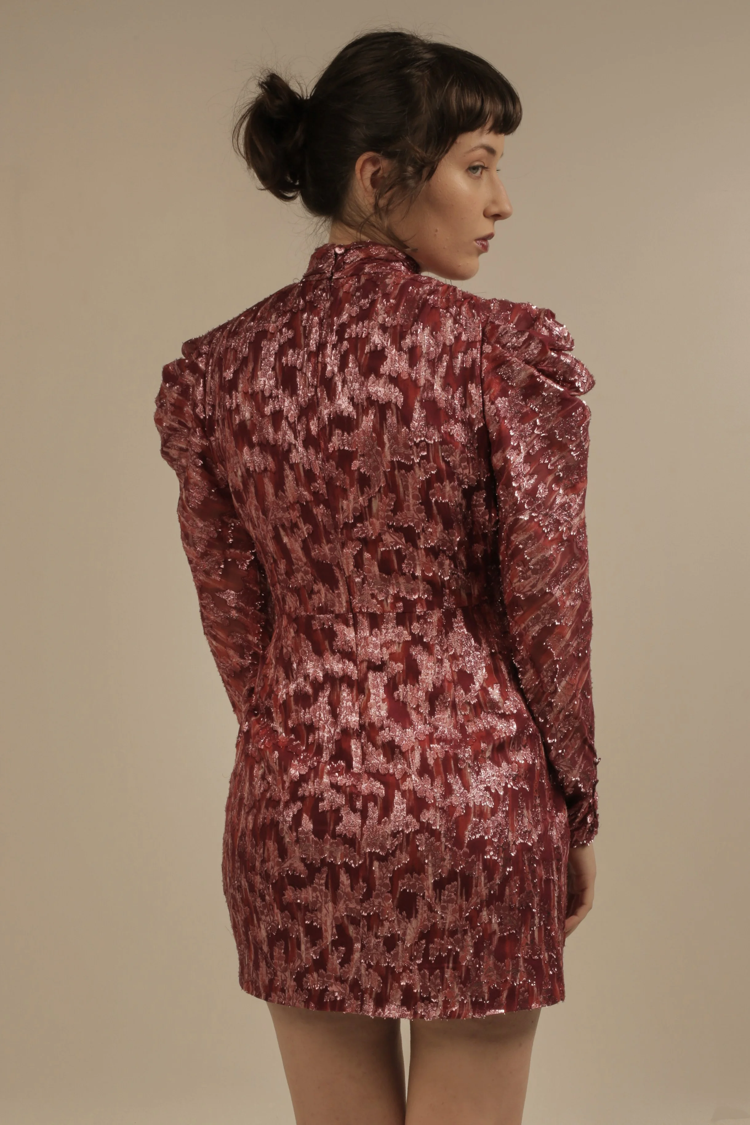 A woman wearing a shiny, patterned red and pink blazer and matching skirt, standing against a plain beige background, with her hair tied in a loose bun.