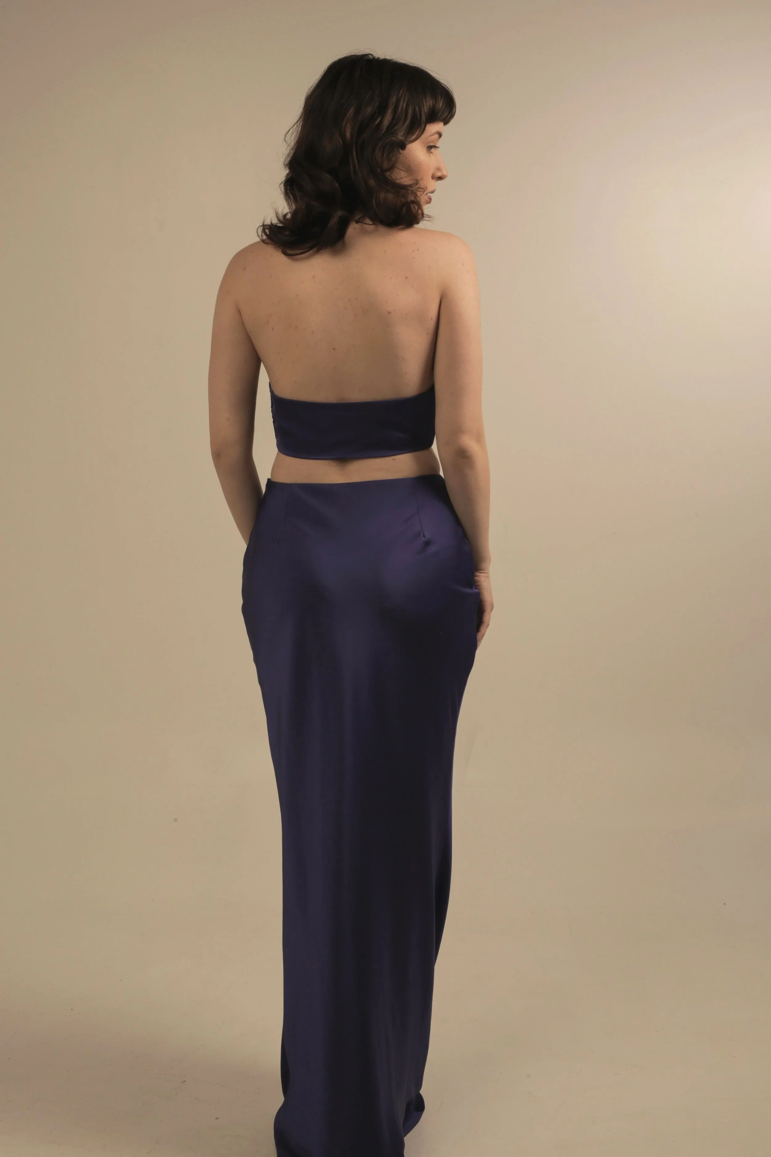 A woman with shoulder-length brown hair and fair skin, standing sideways, wearing a navy strapless top and matching long satin skirt, against a plain beige background.