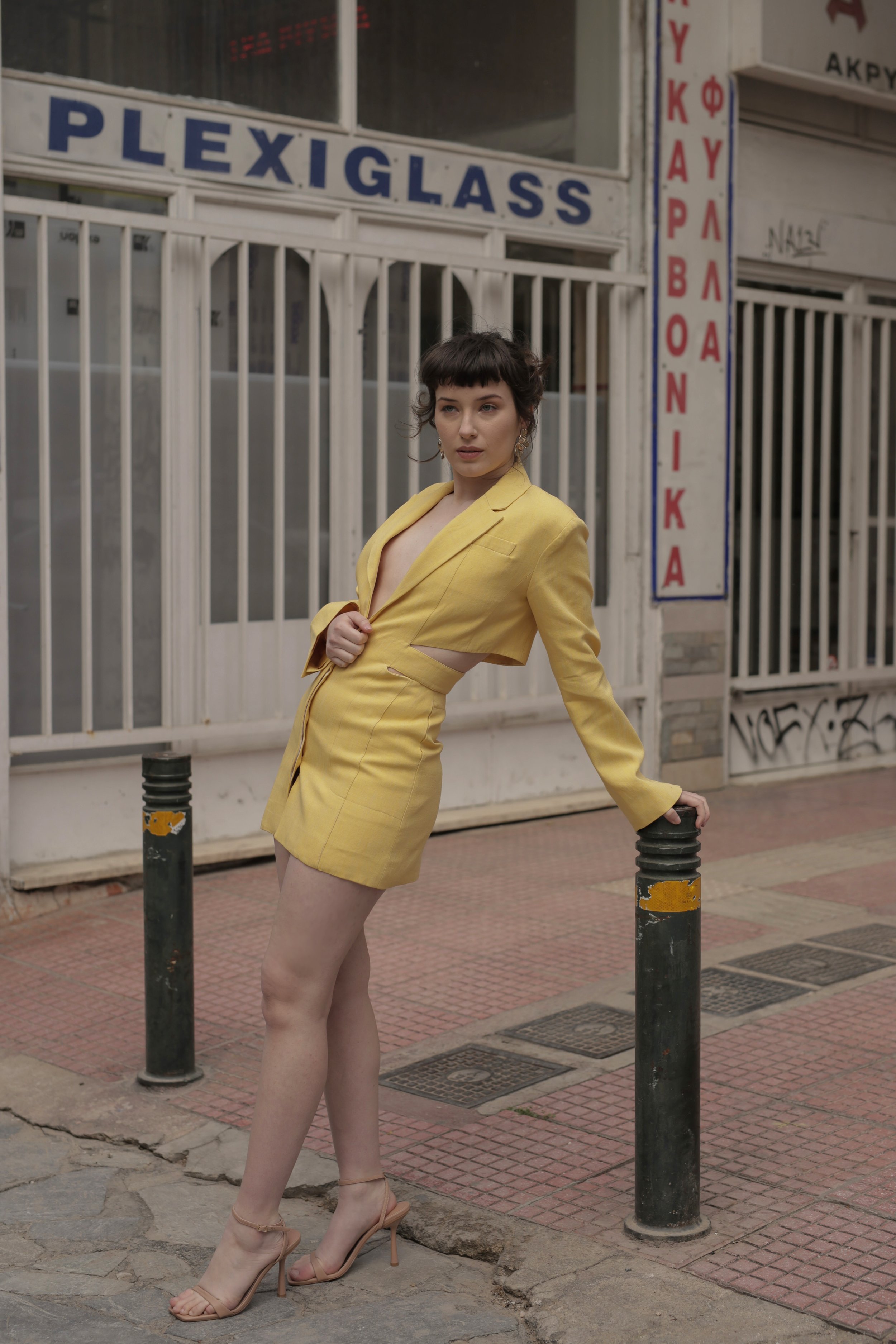 A woman in a yellow blazer dress, standing on a city sidewalk, leaning against a black bollard, with storefronts and graffiti in the background.