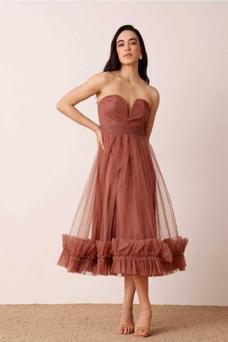 A young woman with long dark hair posing in a strapless, rust-colored midi dress with ruffled hem and sheer fabric, standing against a plain wall.