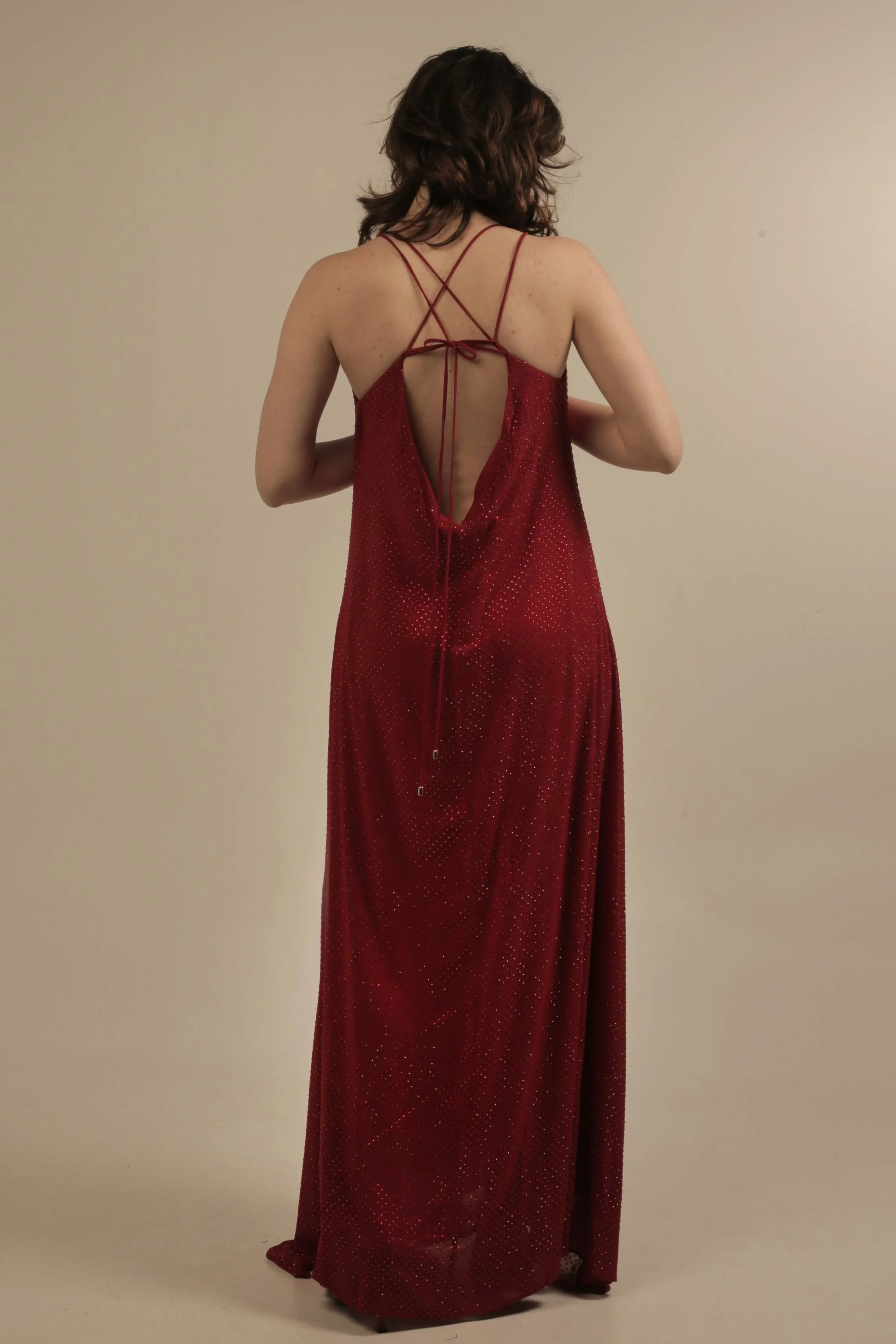 A woman wearing a long, red, sparkly slip dress with a low back and crisscross straps, standing against a plain beige background.