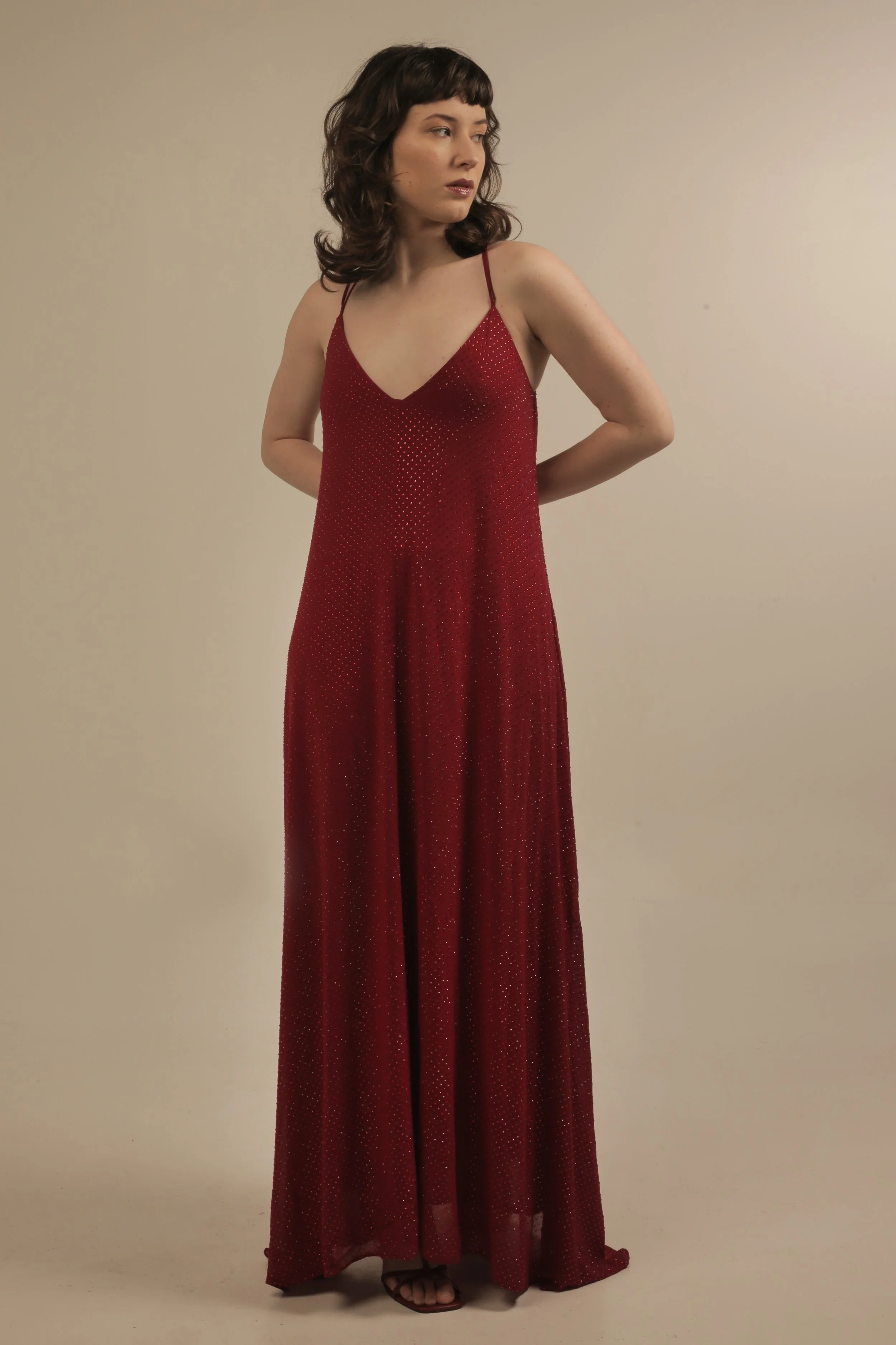 A woman with shoulder-length dark hair, wearing a red, floor-length dress with thin straps and small sparkles, standing against a plain beige background with her hands behind her back.