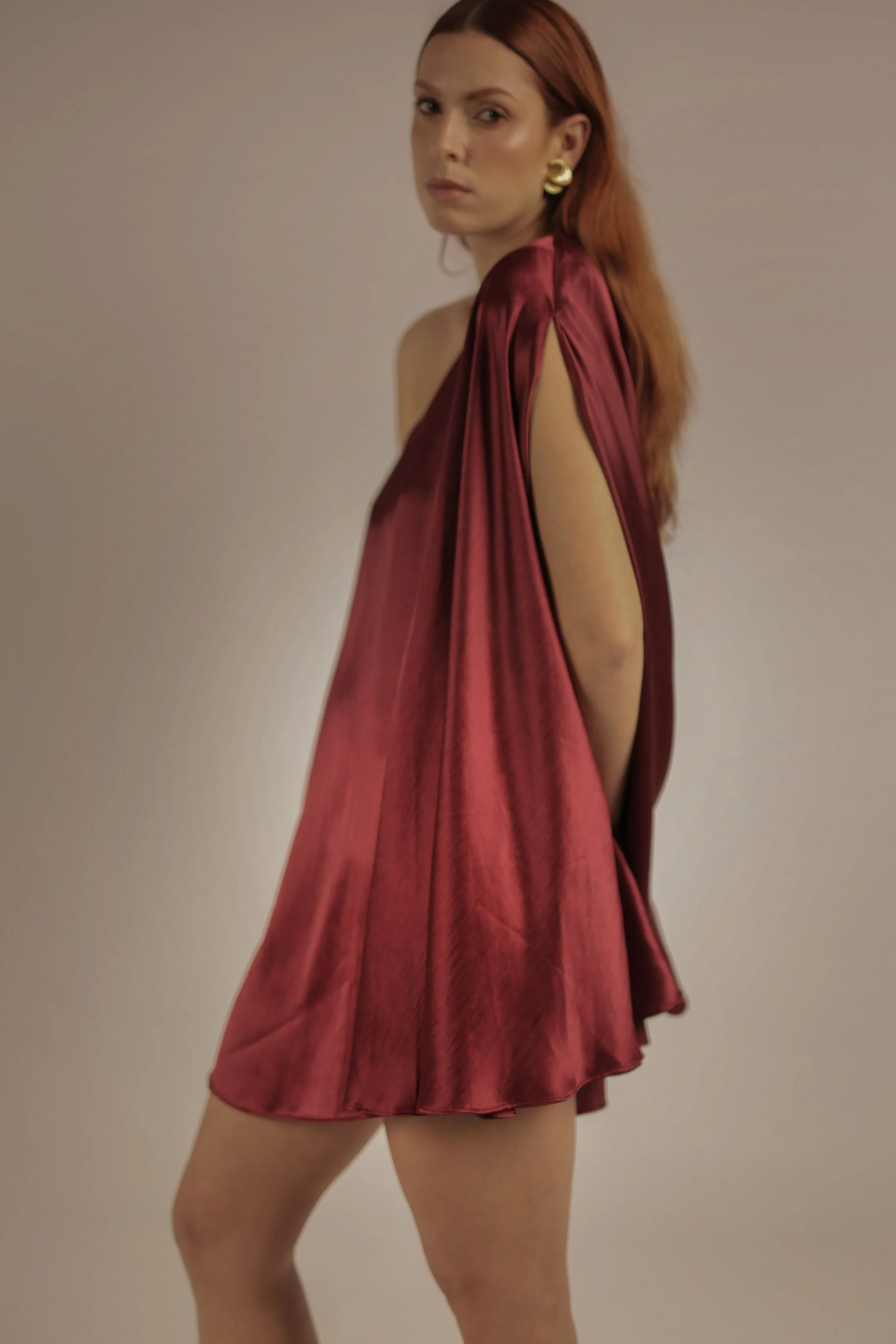 A woman with red hair wearing a shiny, dark red satin dress with open sides and gold earrings, standing against a plain background.