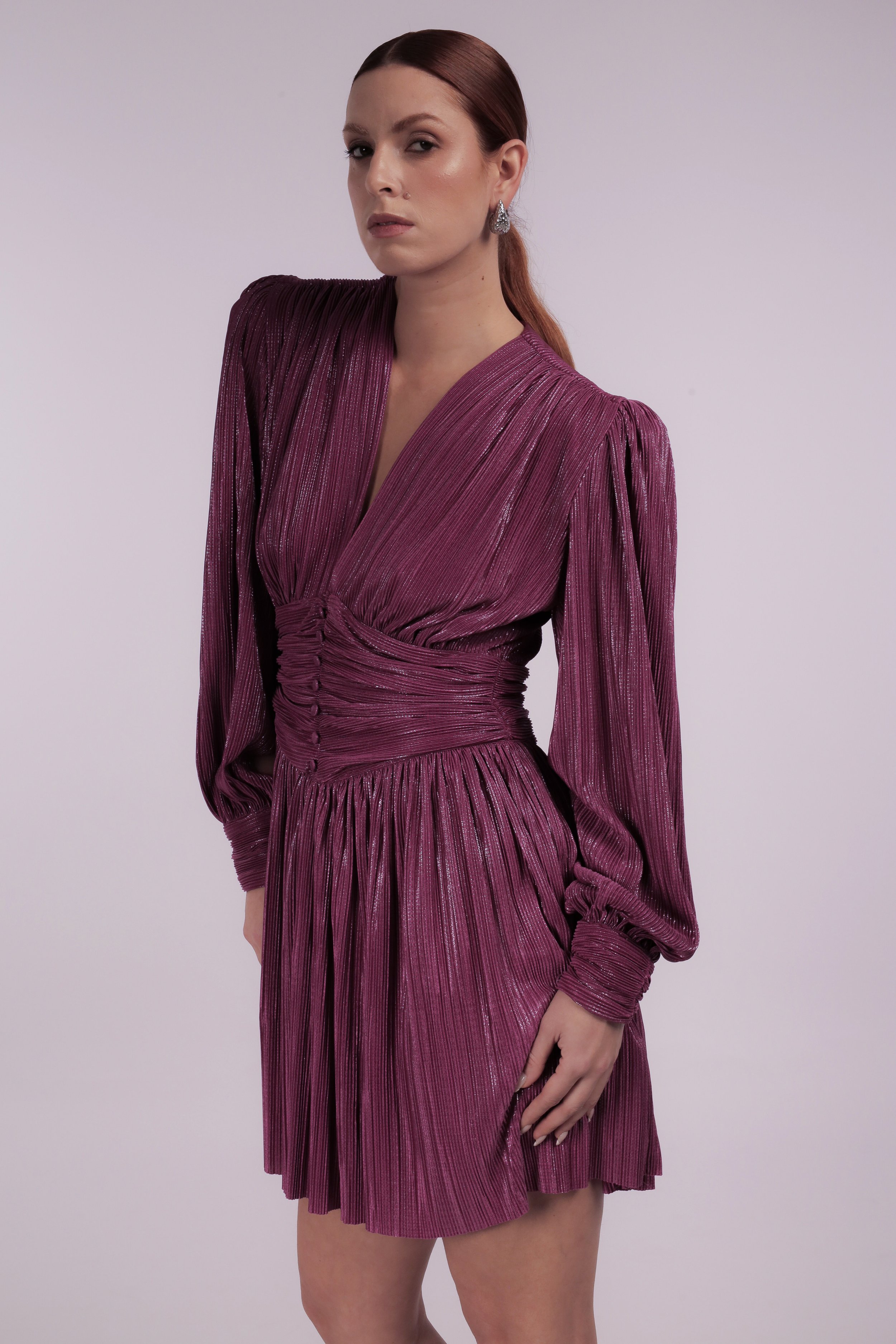 A woman with red hair styled in a ponytail, wearing a shiny, deep pink pleated dress with long puffed sleeves and fitted waist, posing against a plain light background.