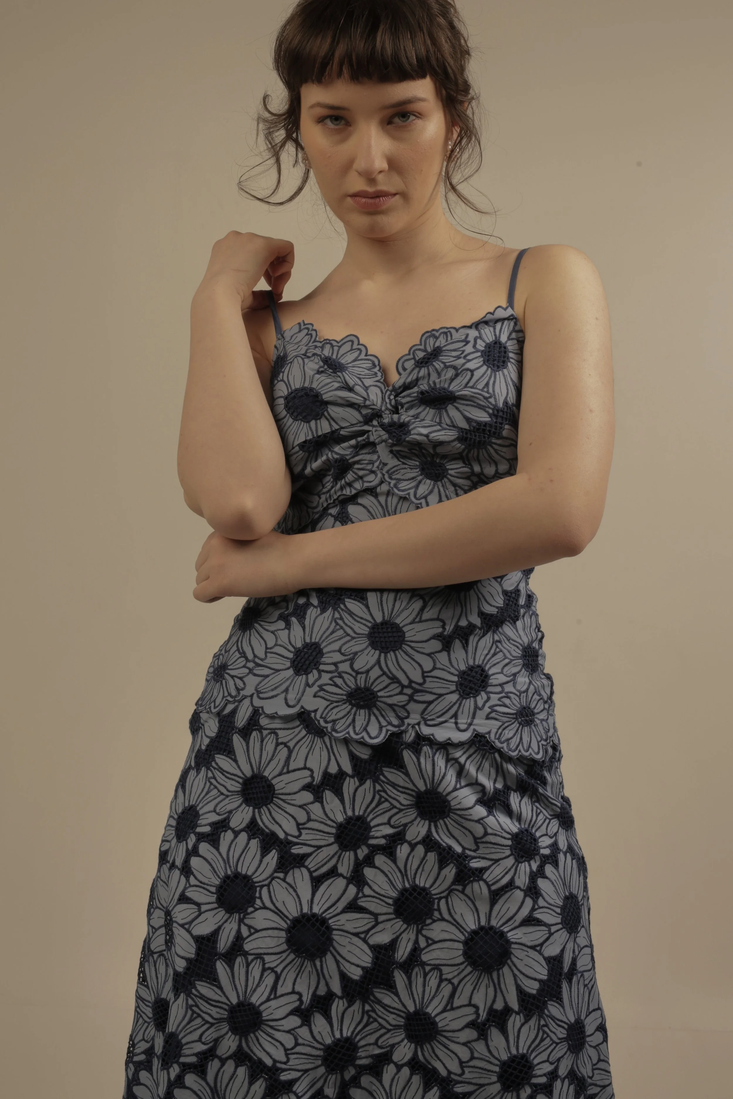A young woman with short brown hair and a serious expression is standing against a plain beige wall. She is wearing a sleeveless, spaghetti strap dress with a black and white daisy pattern. Her right hand is gently touching her left shoulder, and her left arm is crossed over her stomach.