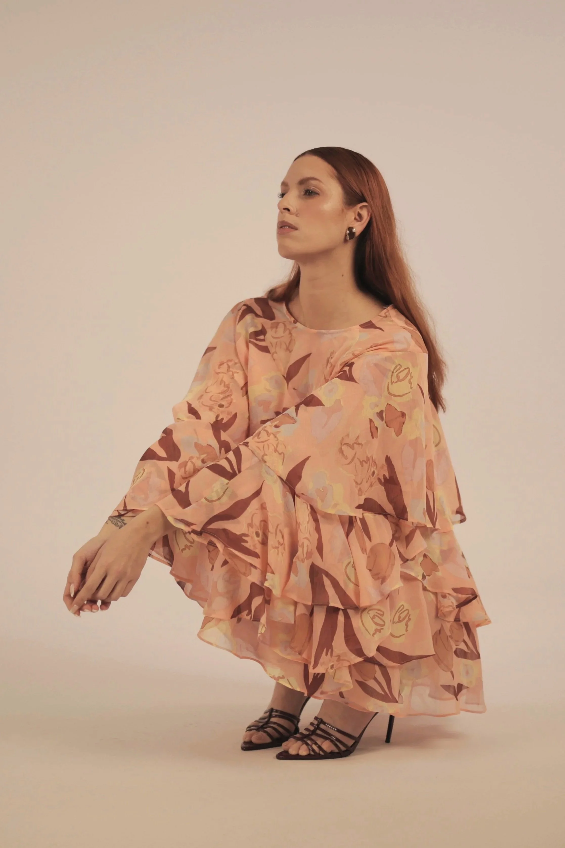 Woman with red hair in a layered floral dress and black strappy heels, seated against a plain background.