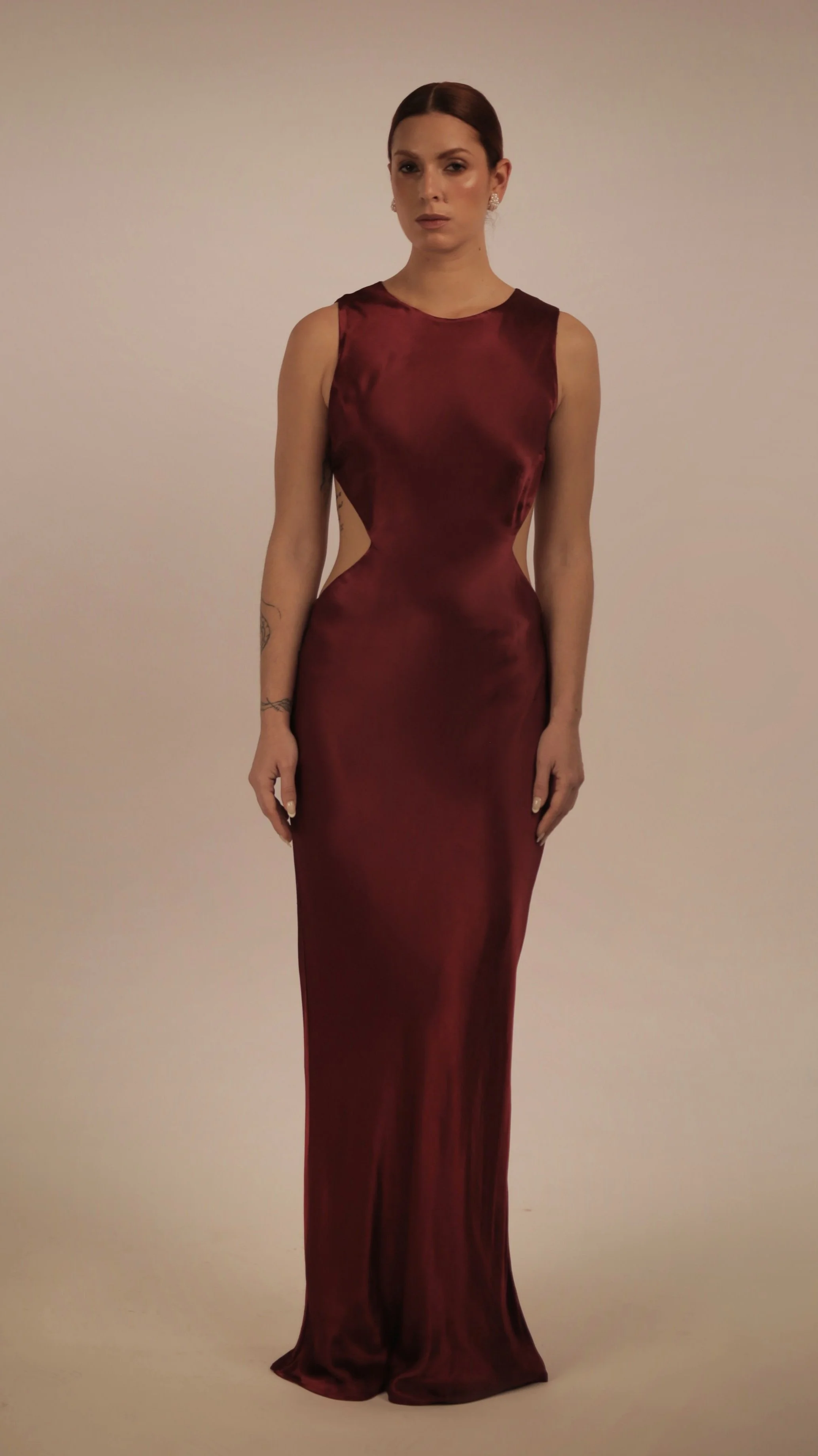 A woman with red hair, wearing earrings, in a long, sleeveless, burgundy satin gown standing against a plain background.