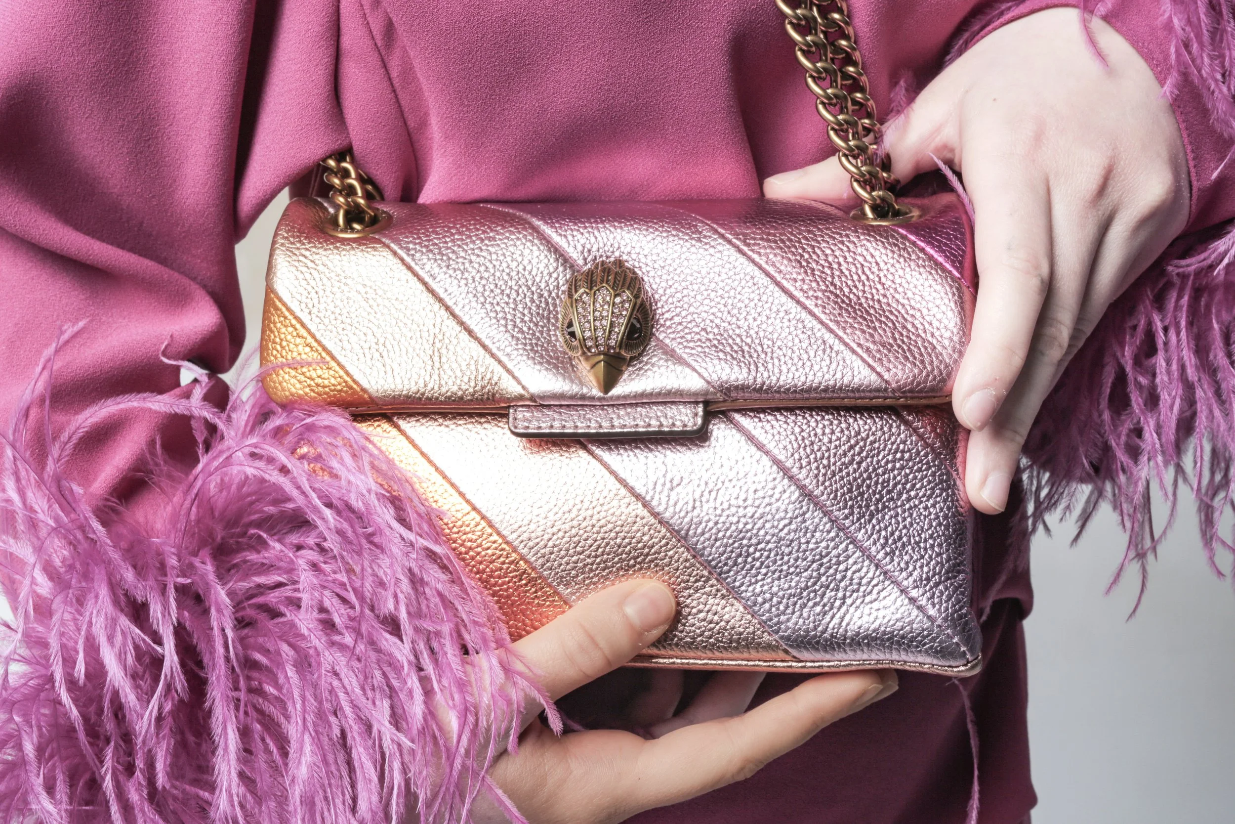 A person is holding a metallic pink clutch purse with a gold chain strap and a decorative clasp, dressed in a pink, feather-trimmed garment.