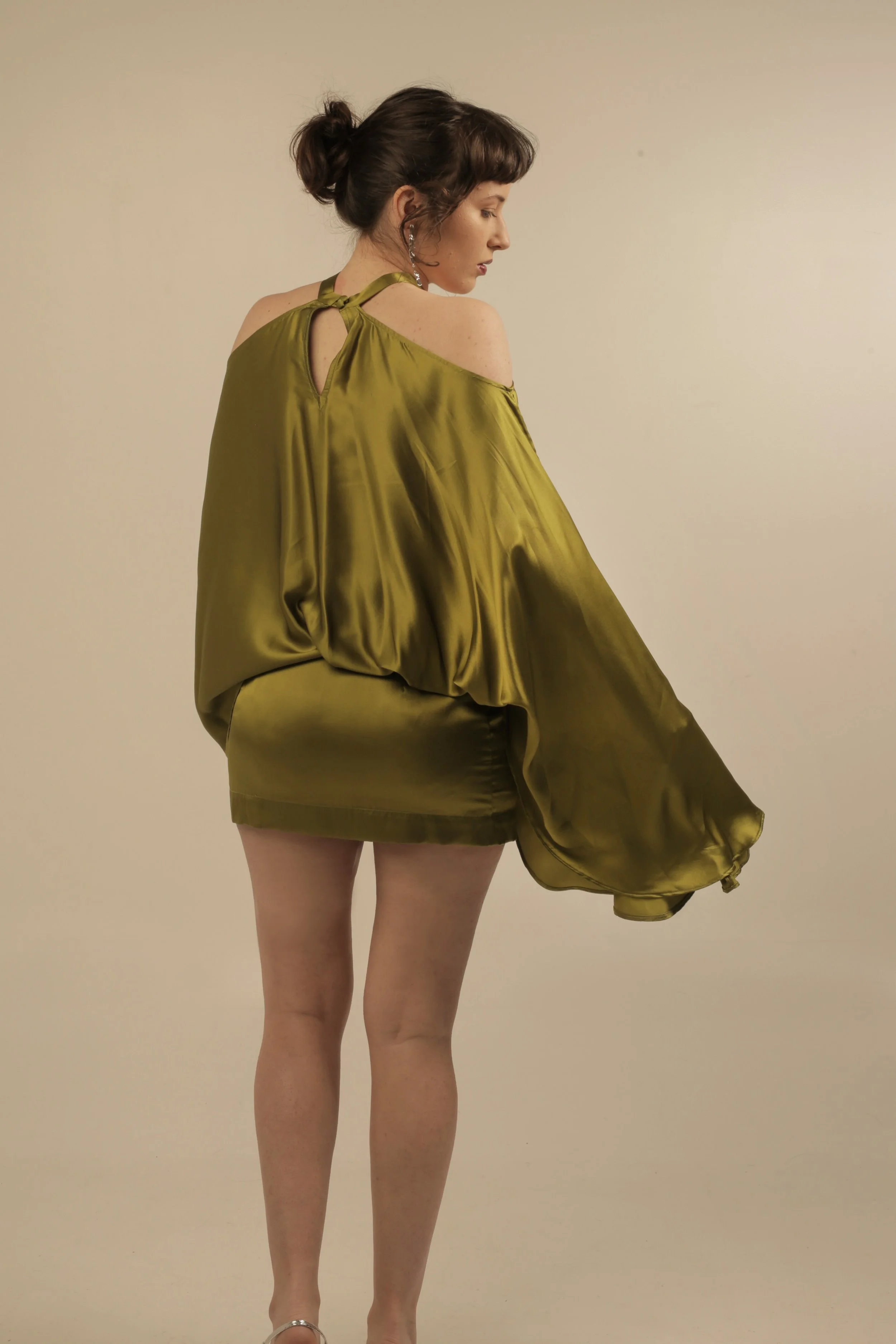 A woman with dark brown hair in a bun, wearing a shiny, olive green, off-shoulder dress with a halter neckline and large earrings, standing sideways against a plain background.