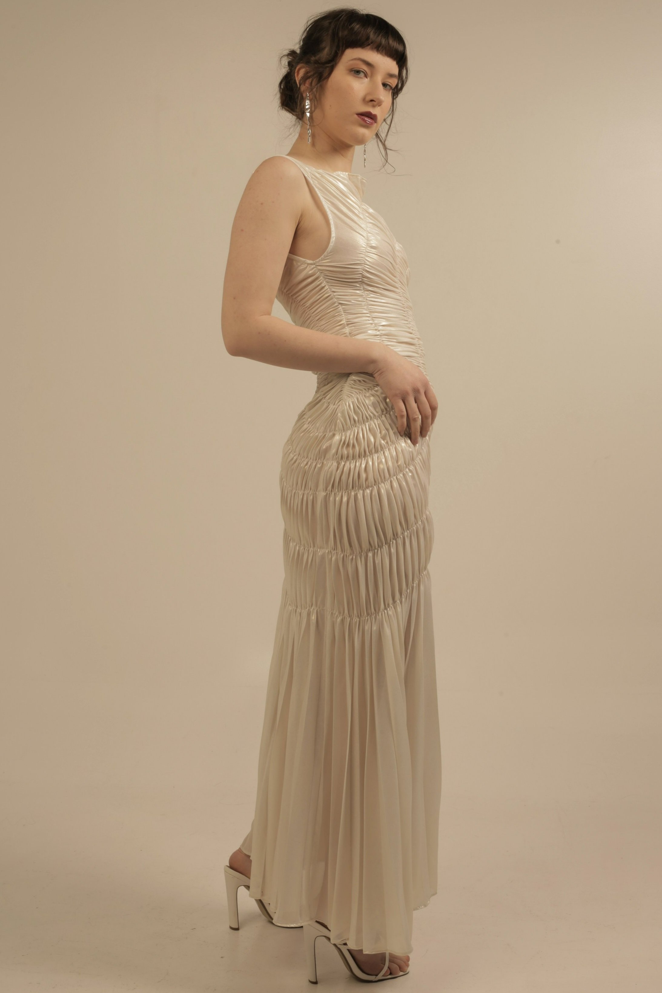 A woman with short dark hair and bangs, wearing a beige, gathered, sleeveless dress with high heels, standing against a plain light-colored background.