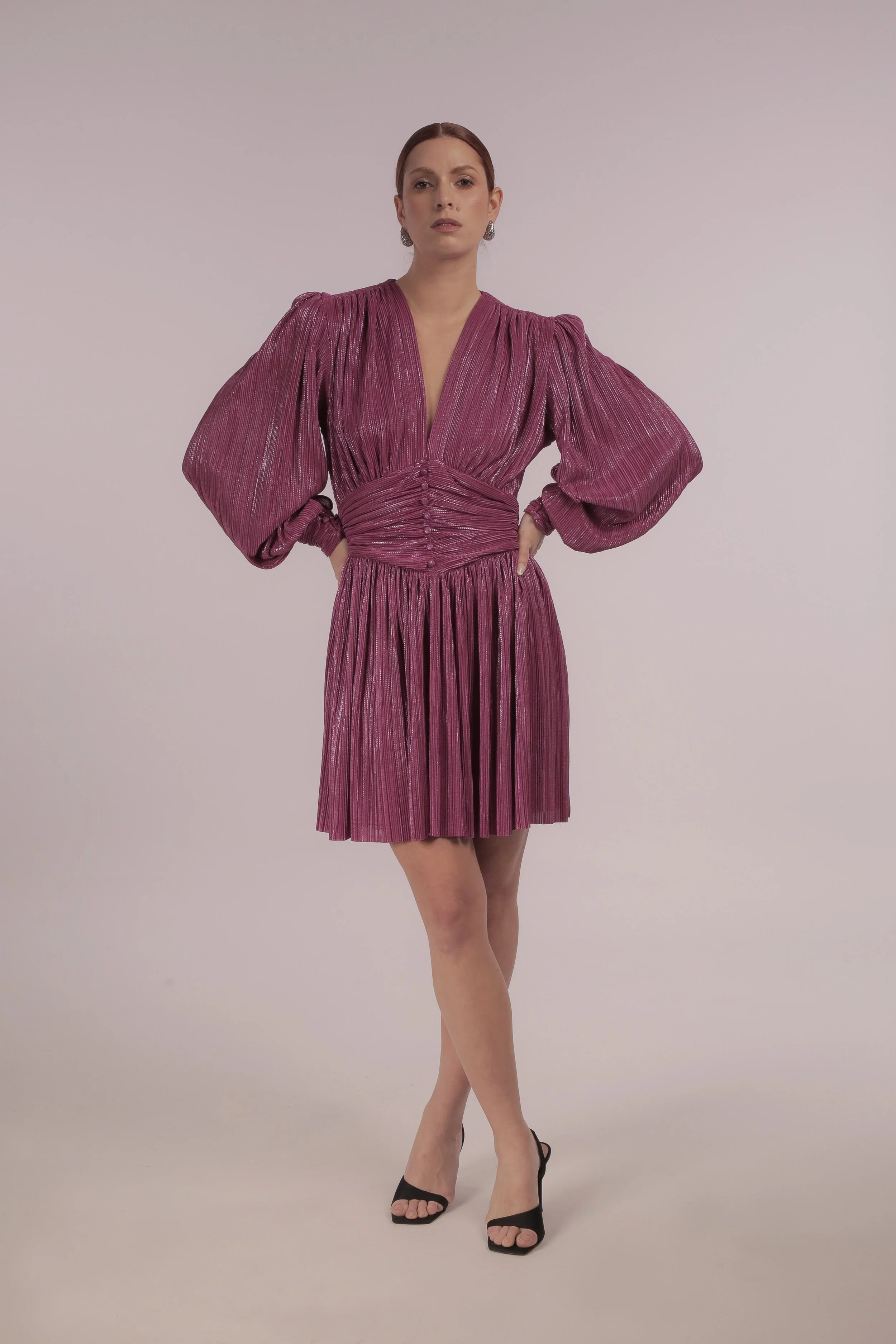 A woman in a shiny, pink, pleated dress with puffed long sleeves and black high heels, standing with her hands on her hips against a plain background.